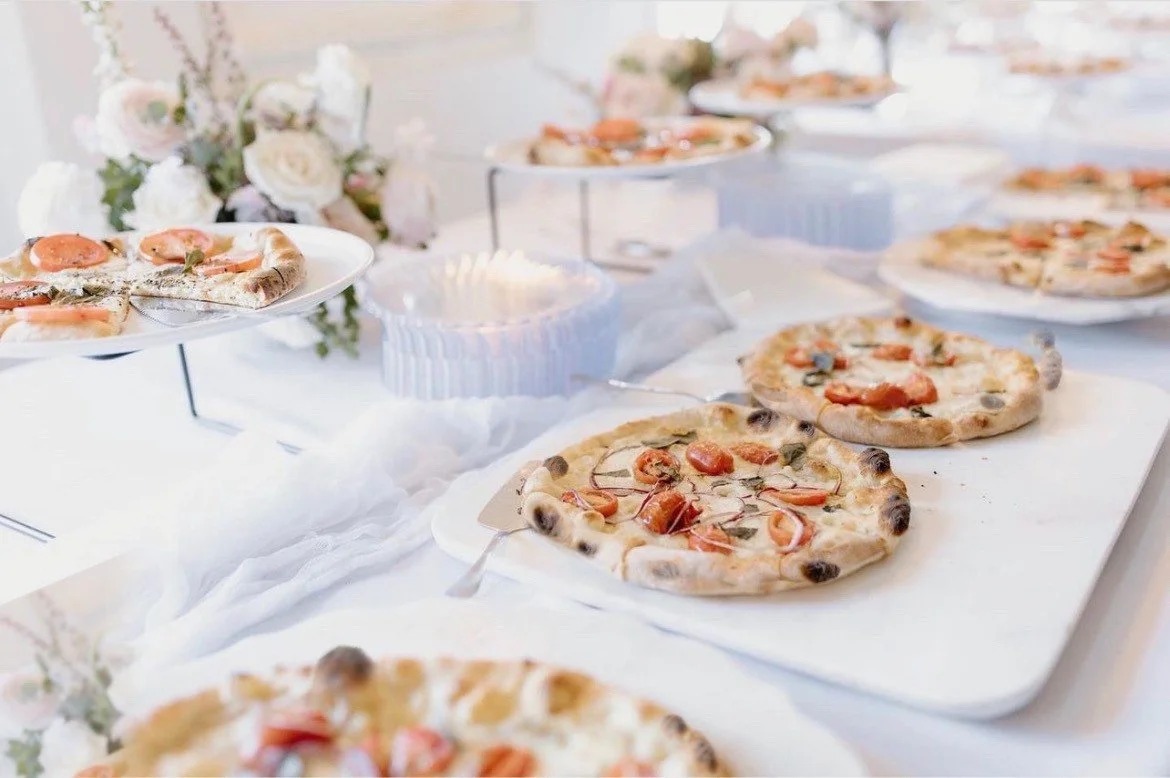 A table set with multiple pizzas, some on a white tray, with floral arrangements in the background, at a celebration or event.