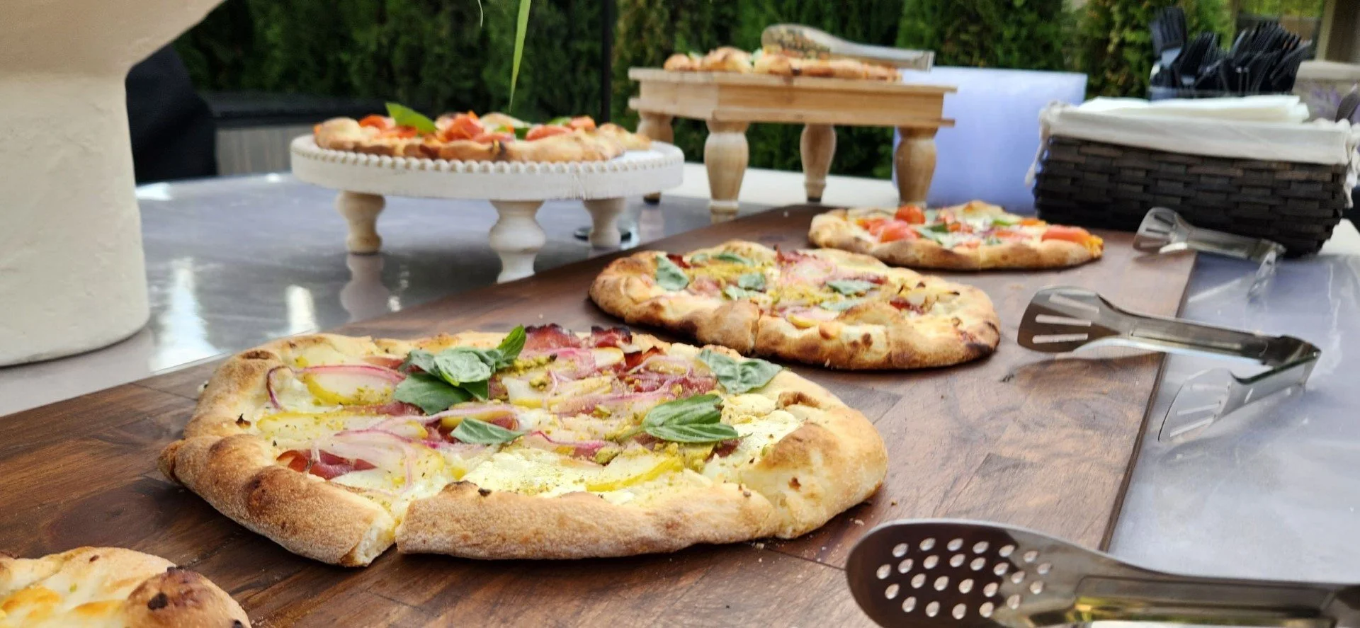Freshly baked pizzas with various toppings on a wooden serving board outdoors, surrounded by tongs and pizza-serving utensils.
