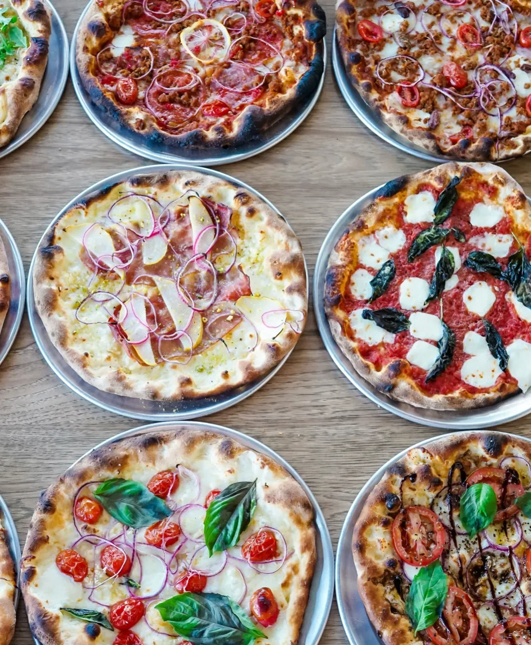 Assorted pizzas with toppings like tomatoes, onions, basil, and cheese on a wooden table.