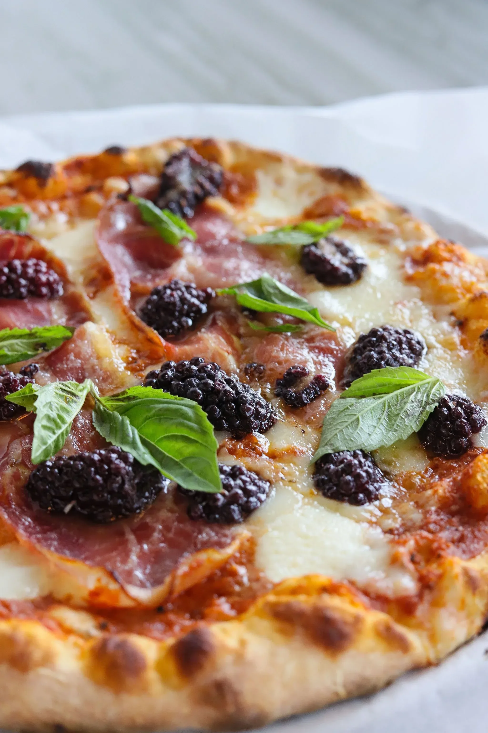 Close-up of a pizza with blackberries, fresh basil leaves, and slices of ham or prosciutto, with a cheesy and tomato sauce base.