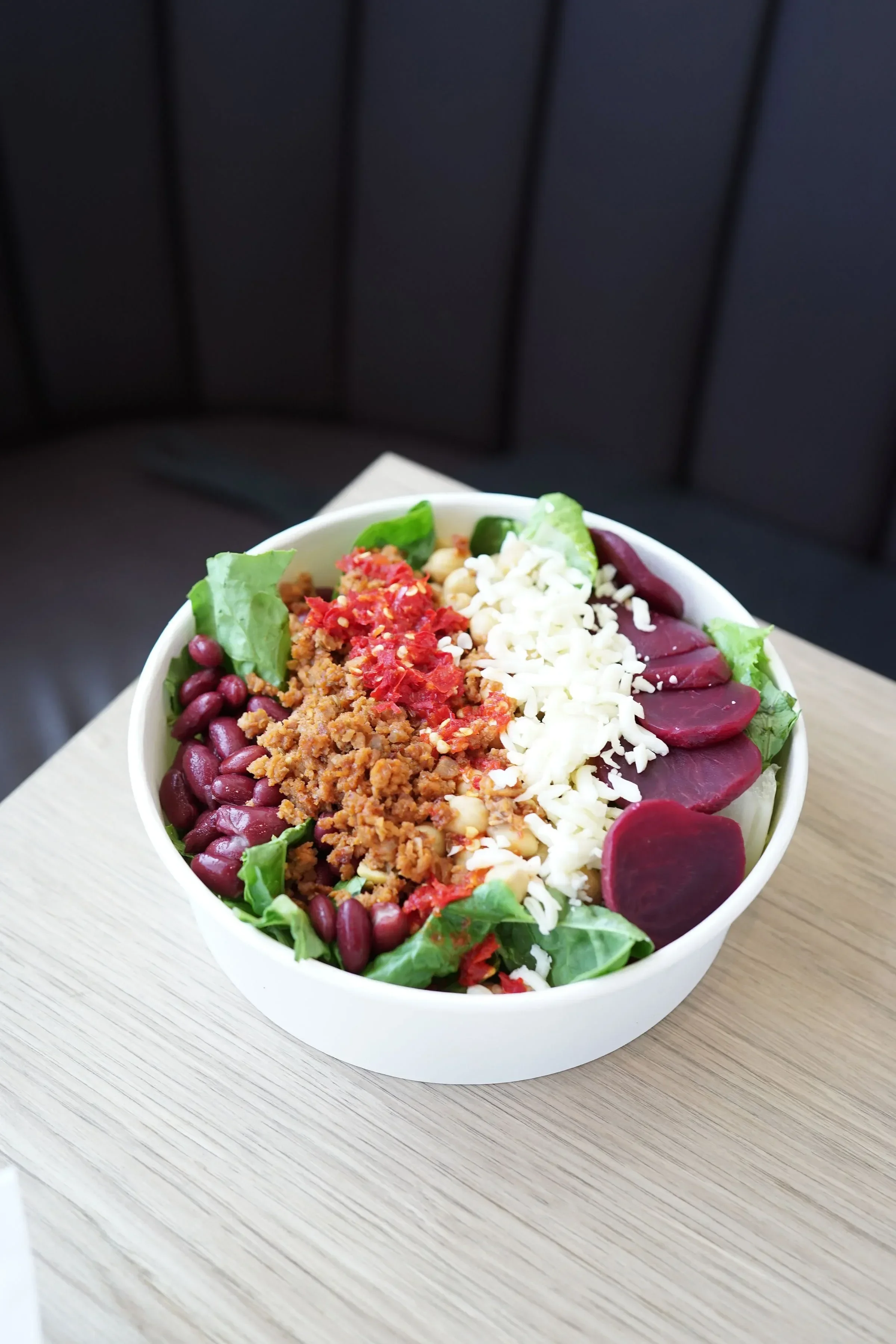 Bowl of salad with lettuce, kidney beans, shredded cheese, ground meat with chili flakes, and sliced beets on a wooden table.