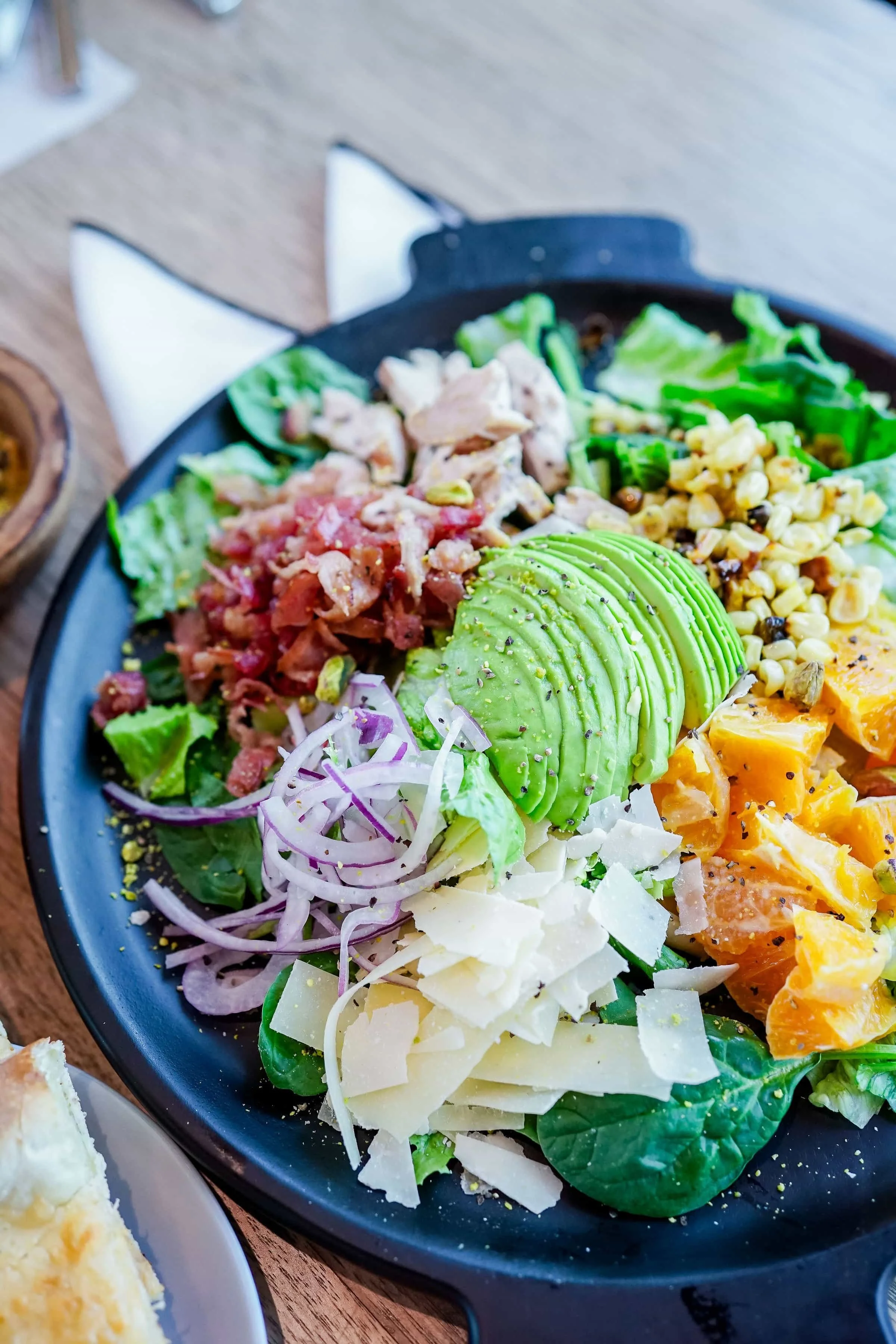 Close-up of a salad in a black bowl with sliced avocado, shredded cheese, chopped tomatoes, lettuce, corn, and other vegetables.