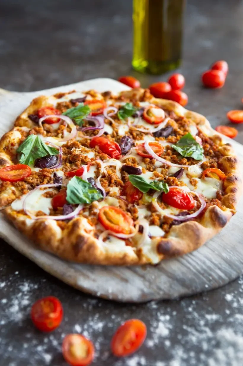 Pizza topped with cherry tomatoes, onions, basil leaves, cooked ground meat, and melted cheese on a rustic wooden board.