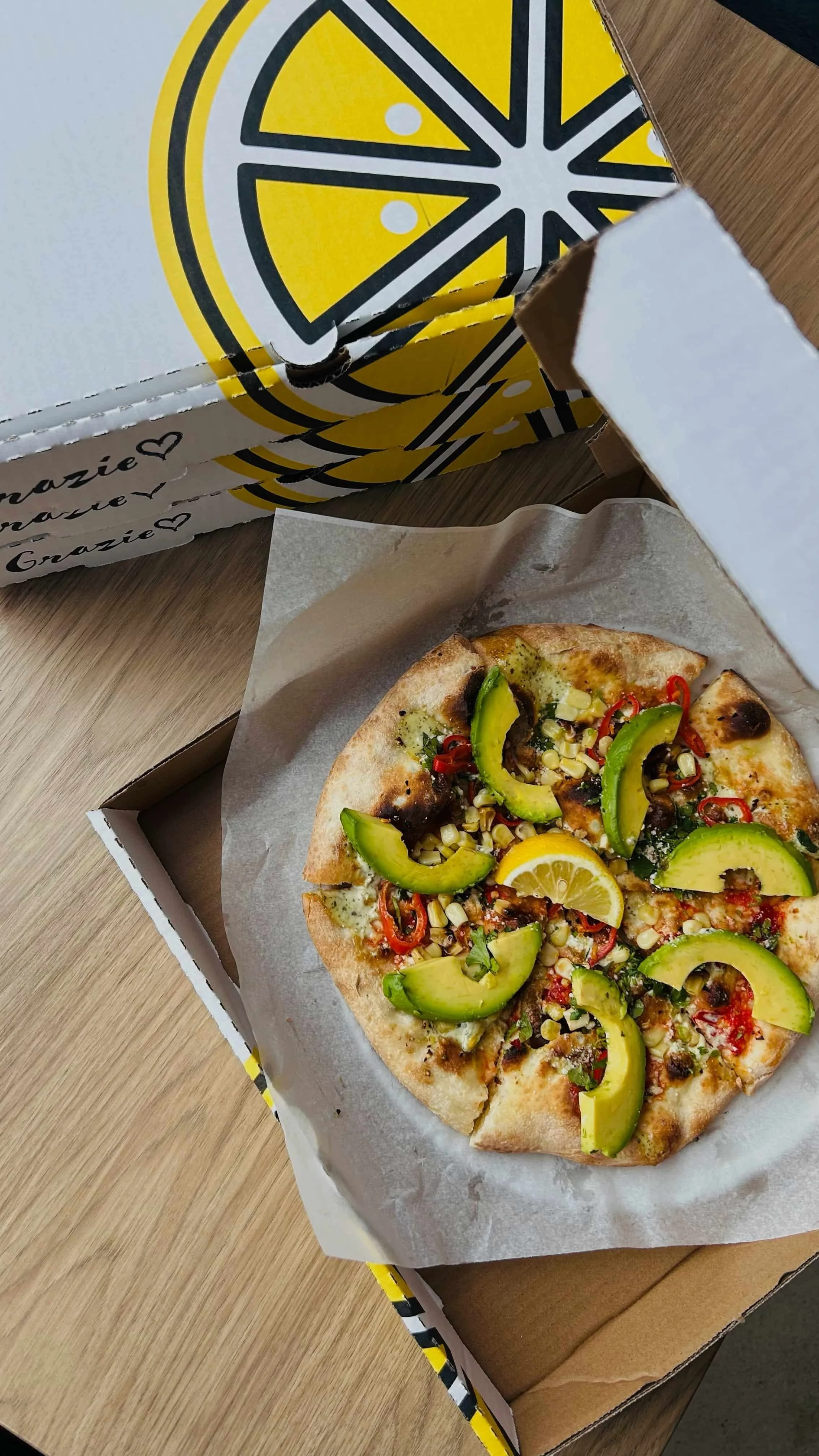 A pizza with avocado slices, corn, red chili peppers, and lemon wedge inside a pizza box on a wooden surface.