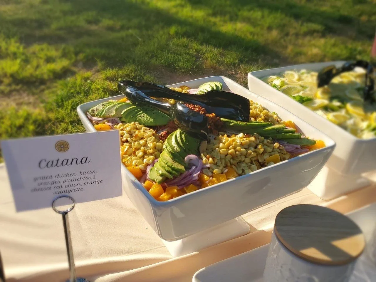 A white bowl of salad with sliced avocado, corn, red onion, and chopped ingredients, with black tongs on top, placed outdoors on a table, with a small sign labeled 'catana' explaining ingredients including grilled chicken, bacon, oranges, pistachios, cheese, red onion, orange vinaigrette.