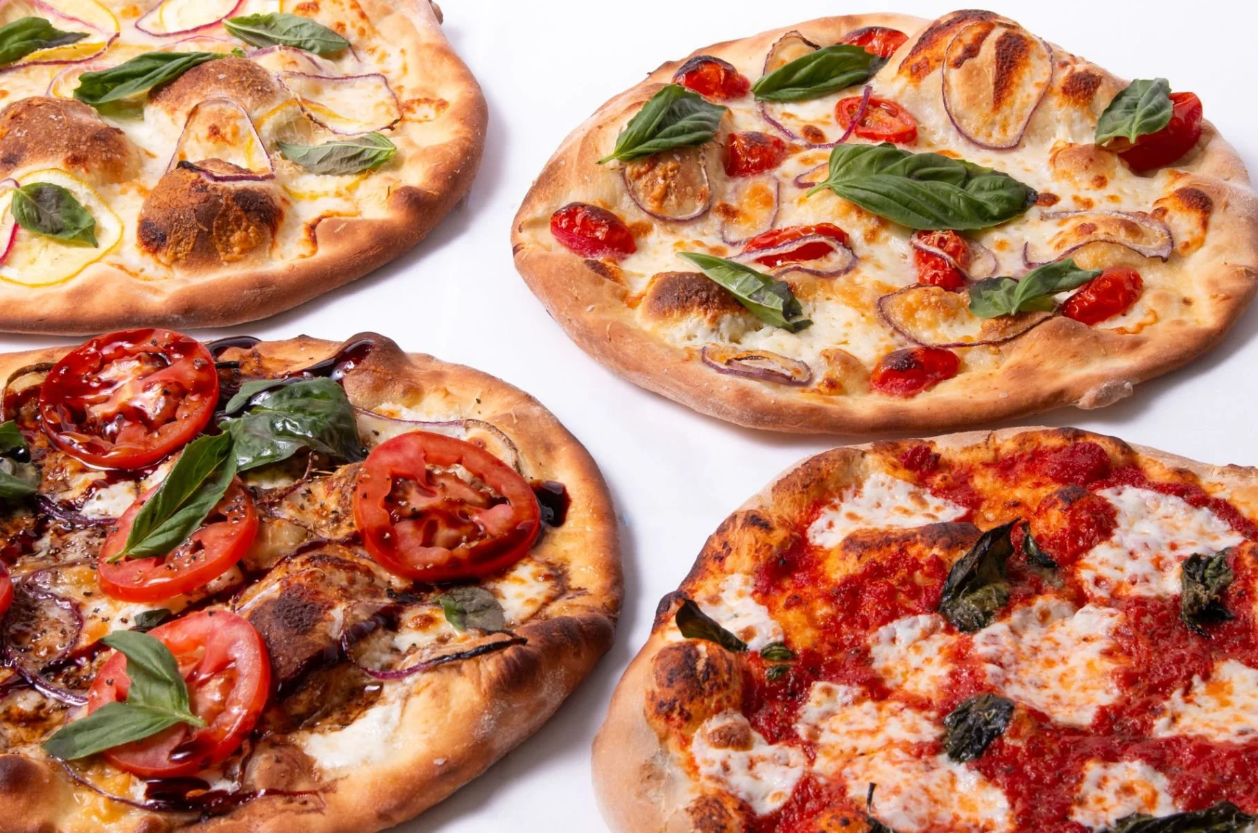 Four different pizzas with various toppings like tomatoes, basil, red onions, and cheese on a white surface.