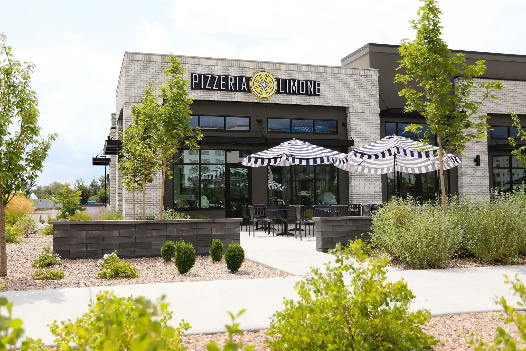 A modern pizzeria named Pizzeria Limone with outdoor seating and striped umbrellas, surrounded by greenery.