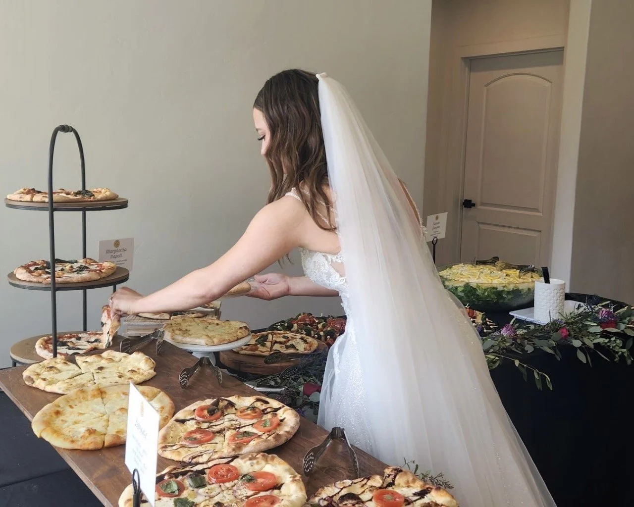 Bride in white wedding dress and veil serving herself pizza at a buffet table with various pizza options in a banquet hall.