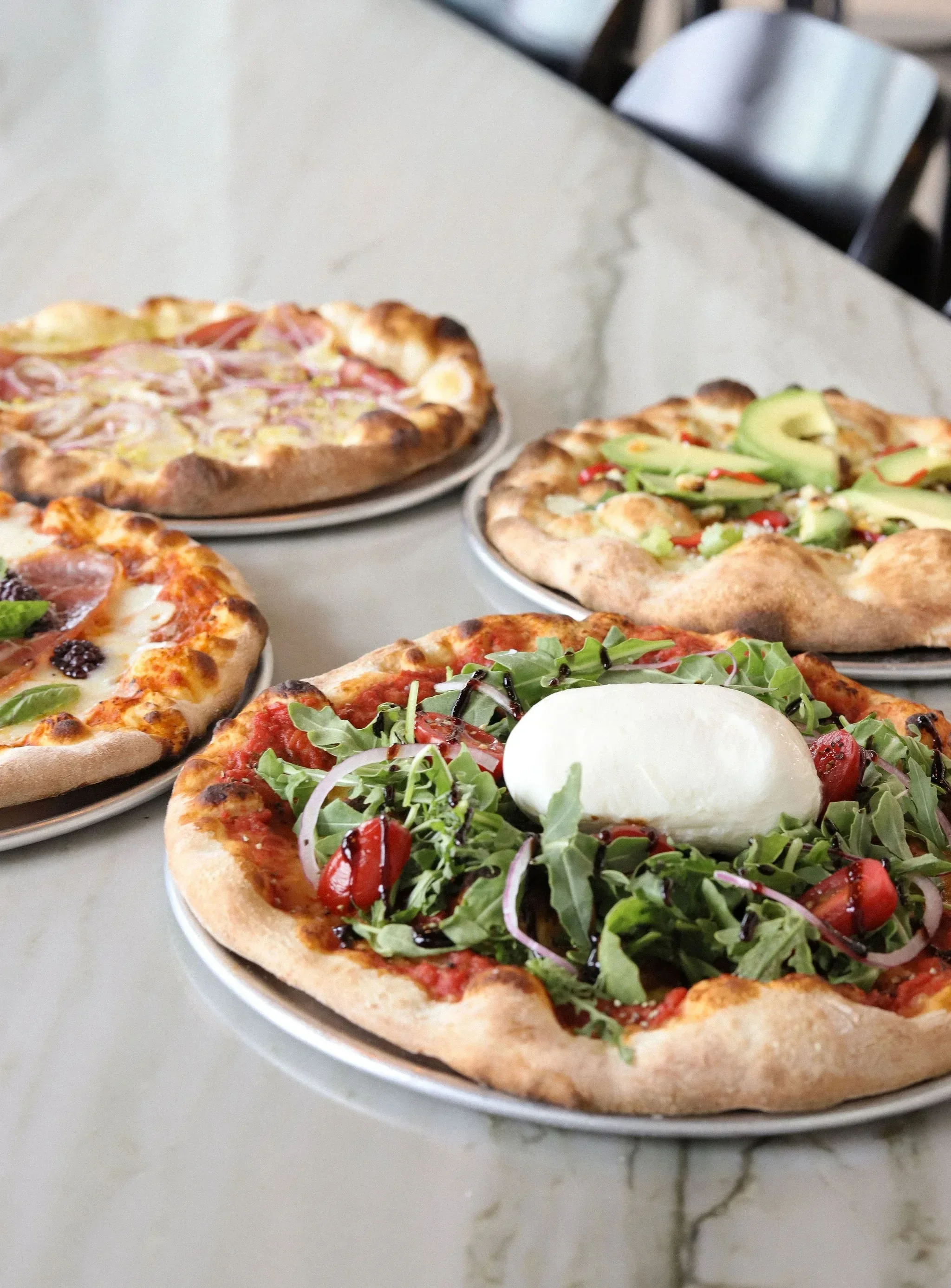 Three pizzas on plates on a table, with one pizza topped with arugula, cherry tomatoes, and mozzarella, and the other with various meats and vegetables.