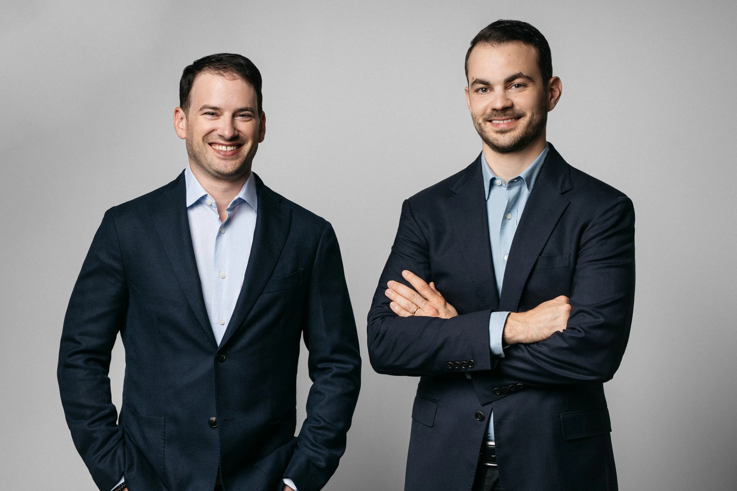 Two men in suits standing against a plain gray background, smiling and posing for the photo. The man on the left has his hand in his pocket, and the man on the right has his arms crossed.