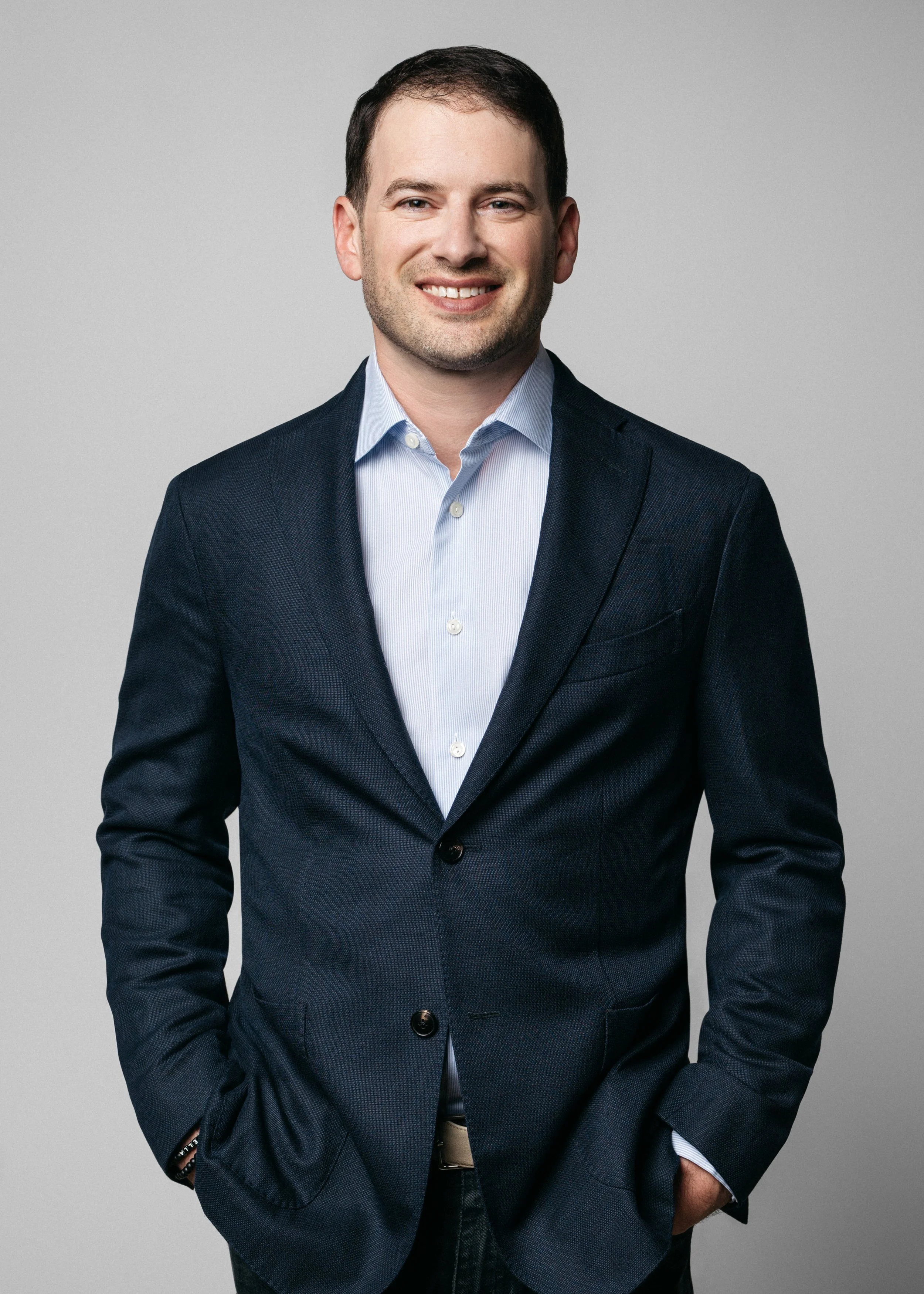 A man wearing a dark suit and light blue shirt, smiling with hands in pockets, against a plain gray background.