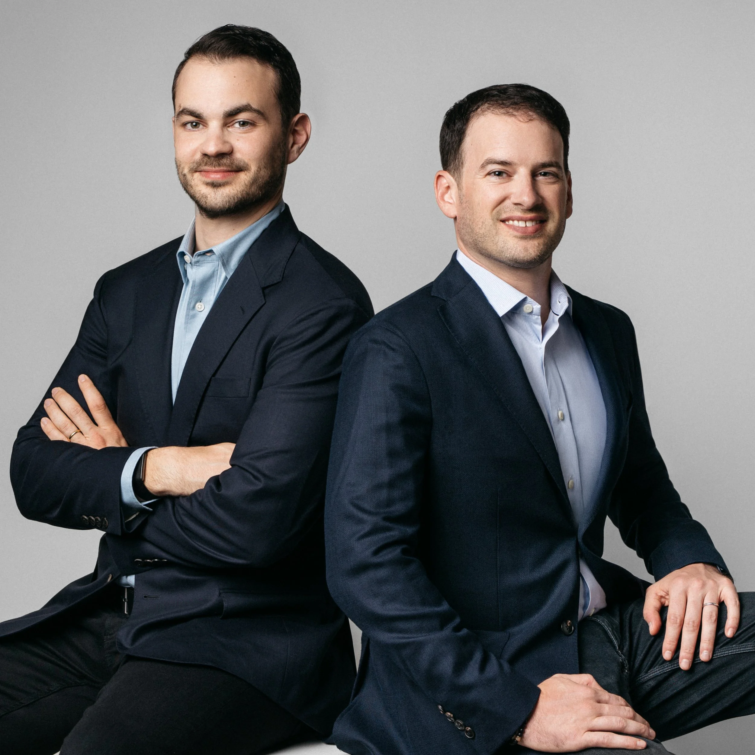 Two professional men in suits sitting back to back, posing for a corporate photo against a plain gray background.