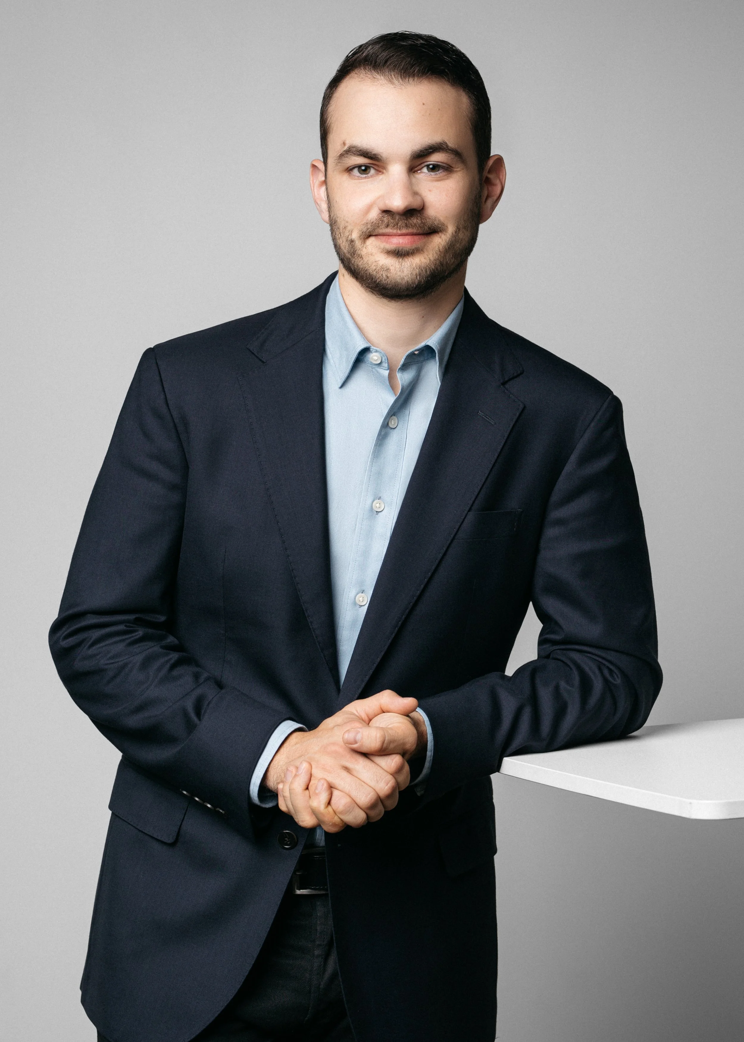 Professional man in a dark suit and light blue shirt standing with hands clasped in front of a white table against a light gray background.