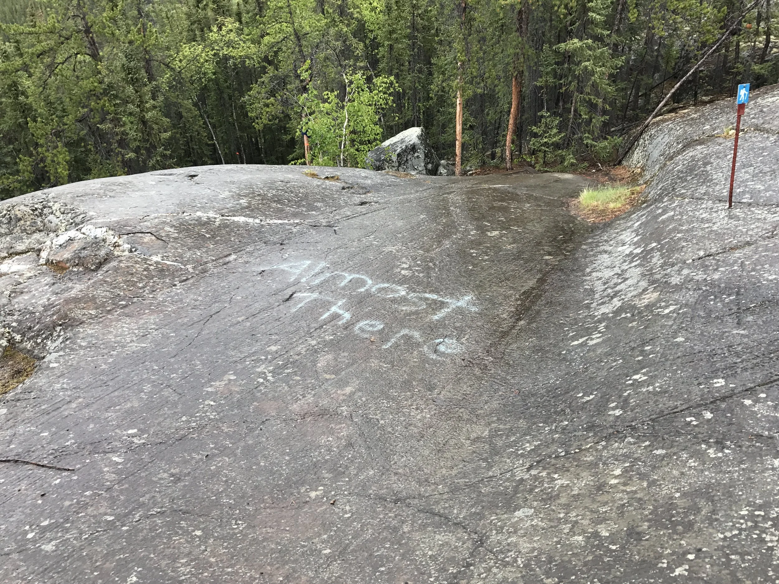 A large, flat rock surface with a painted message that reads 'Almost There' in white chalk, surrounded by a forest with trees in the background, and a small trail or pathway nearby.