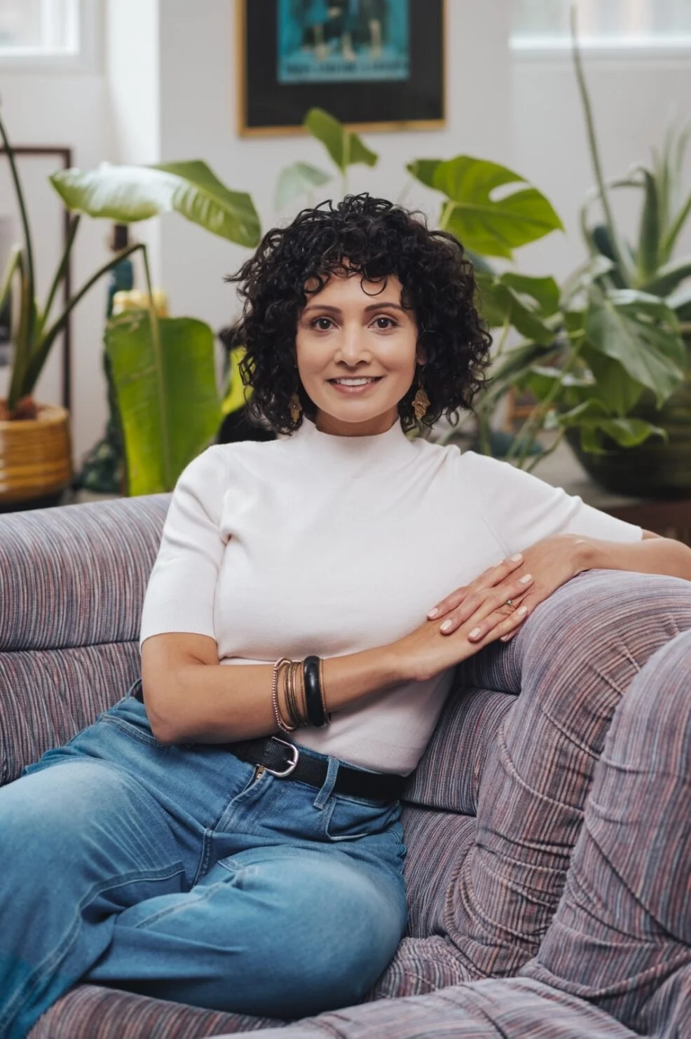 Kathleen Sydney, a Psychotherapist in Toronto is pictured sitting down on a sofa wearing a white top and blue jeans with green plants and framed pictures on the wall.