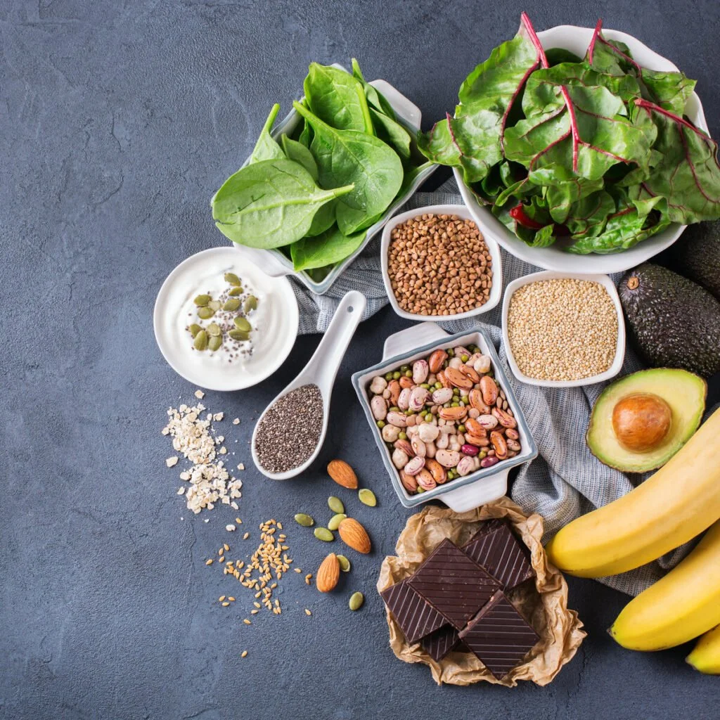 Assorted fresh fruits and vegetables, seeds, nuts, grains, dark chocolate, and oats arranged on a dark surface.