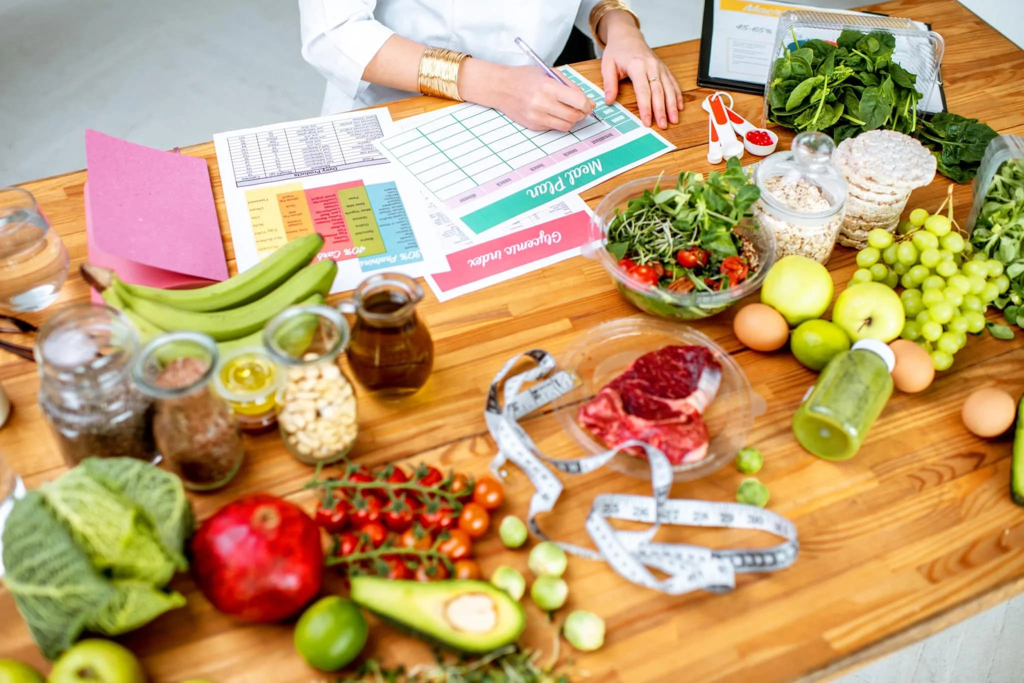 A person working at a wooden table with fresh vegetables, fruits, meat, grains, and health tracking documents, possibly for meal planning or health analysis.