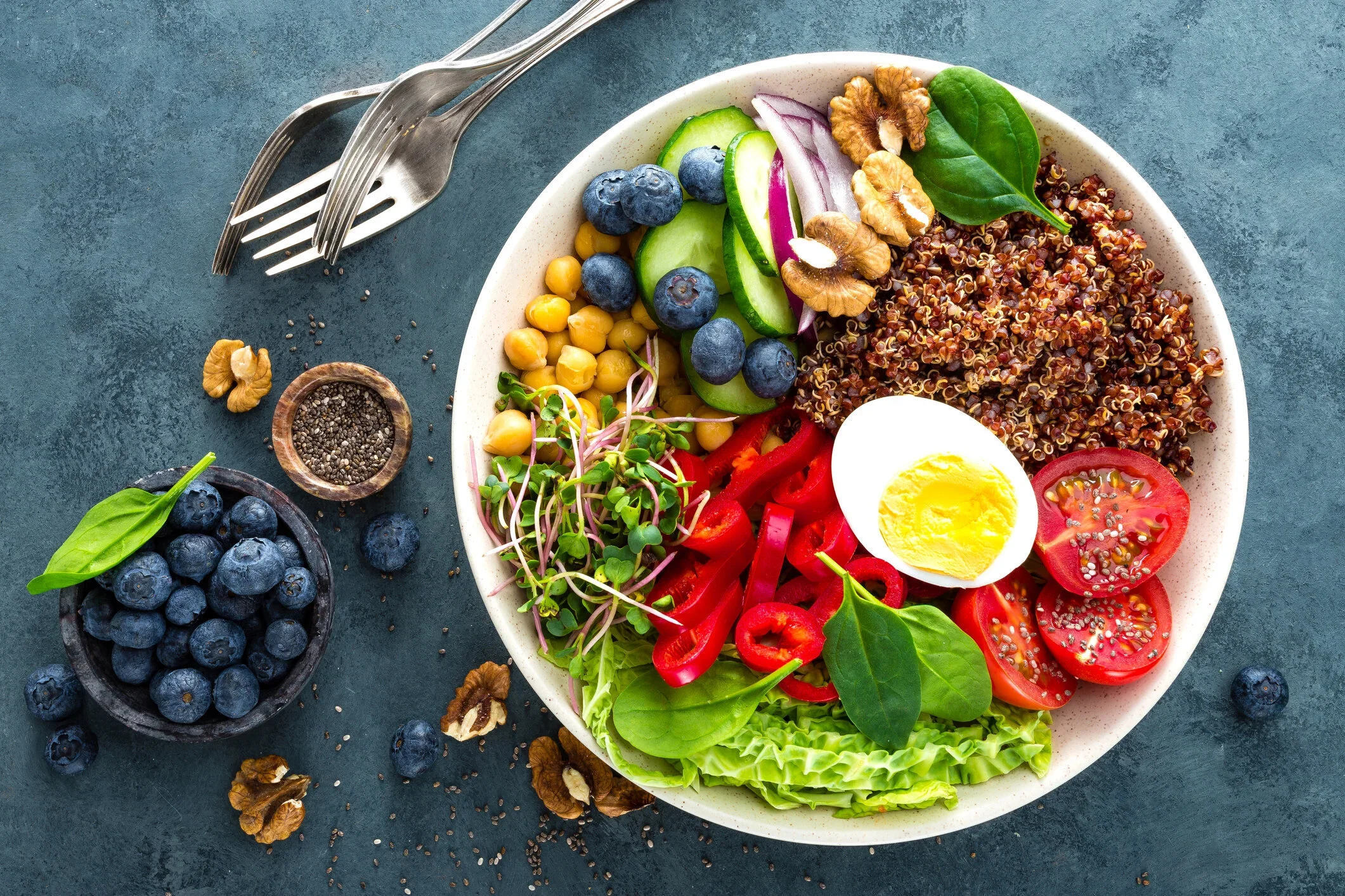 A colorful salad bowl with cherry tomatoes, a halved boiled egg, red bell peppers, cucumbers, blueberries, chickpeas, microgreens, spinach leaves, and quinoa on a dark textured surface, with a spoon and fork nearby.
