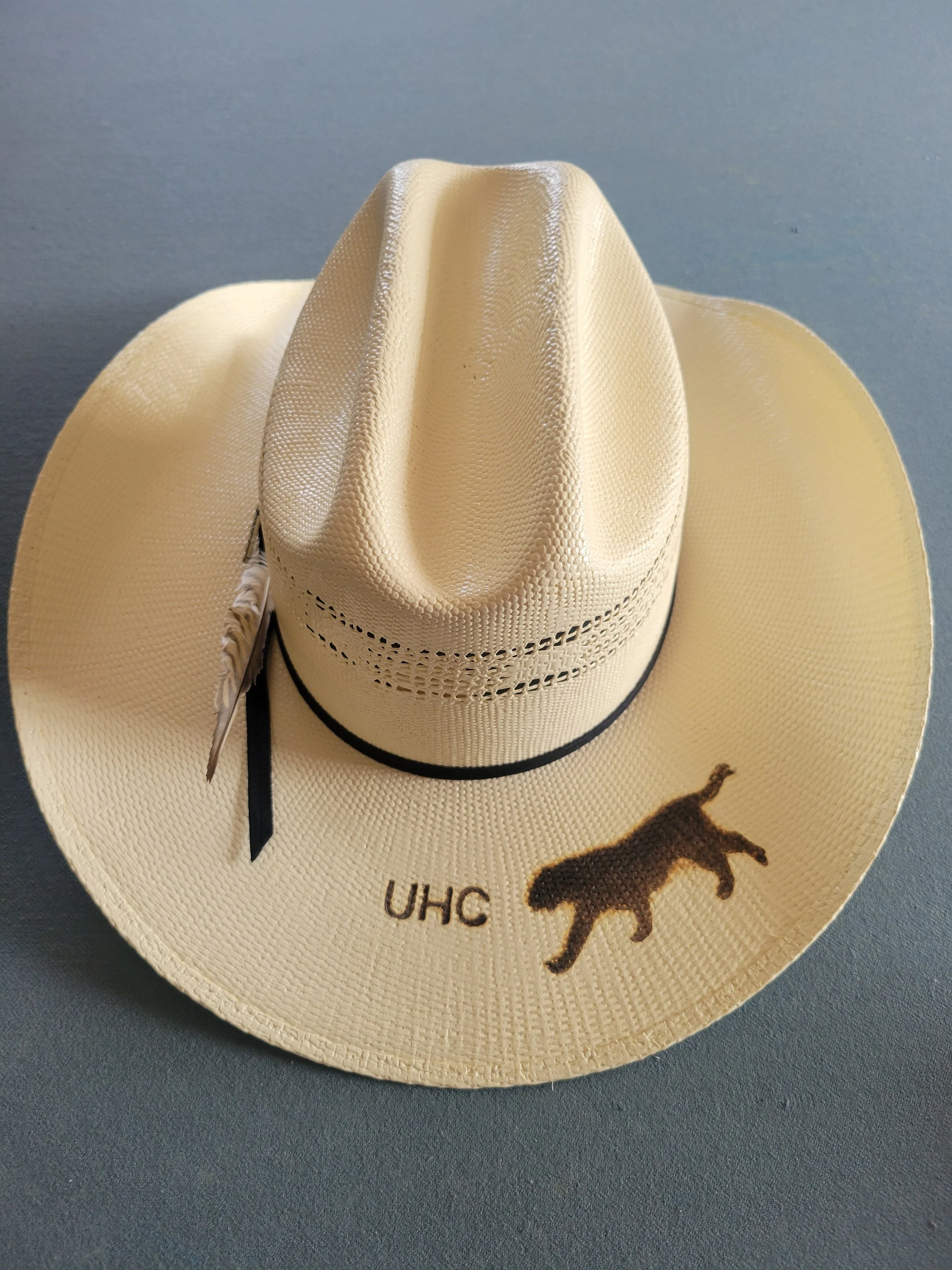 Cream-colored cowboy hat with a black band, a feather, and embroidered silhouette of a wolf and the letters "UHC".