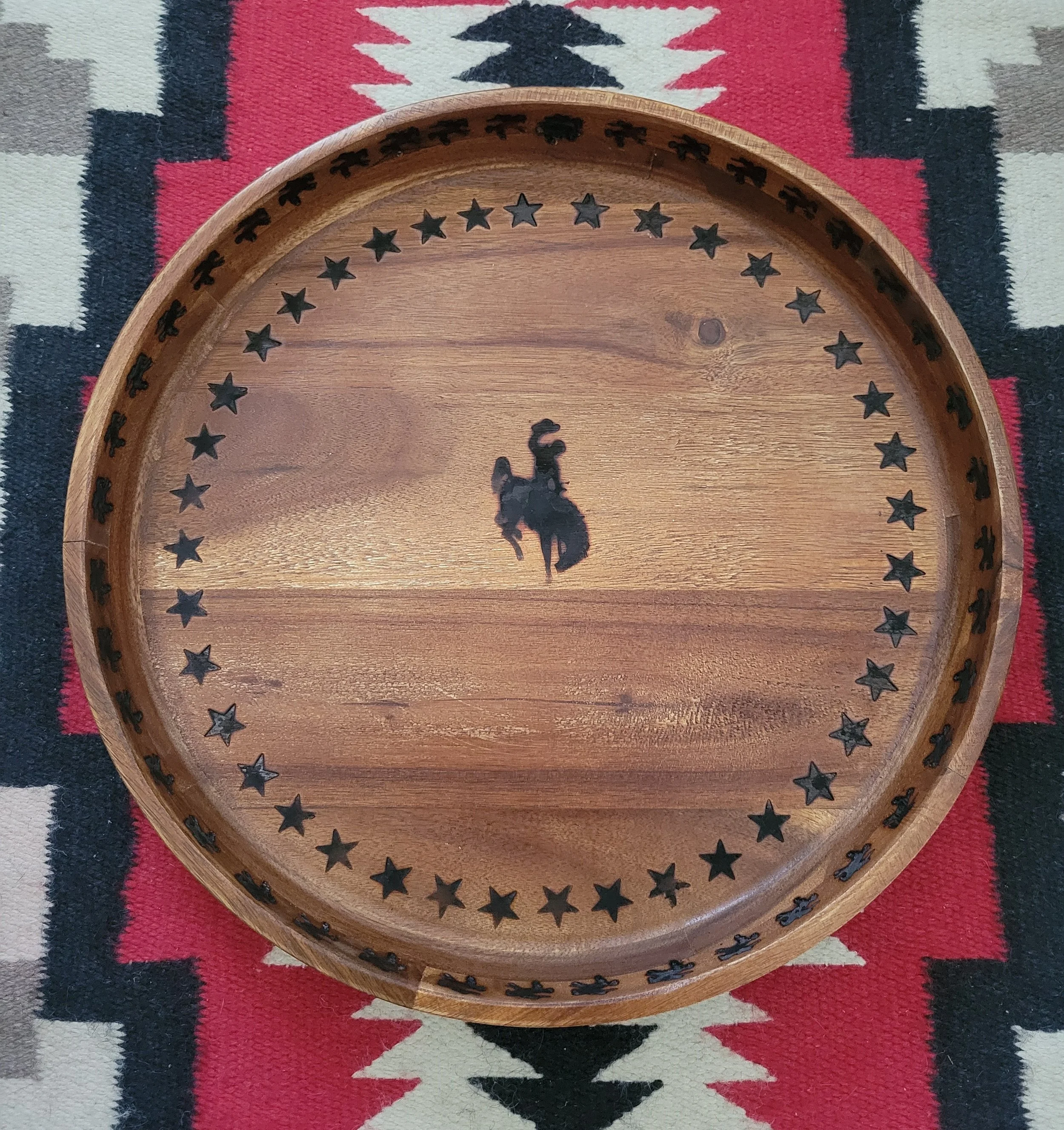 A round wooden tray with a silhouette of a cowboy on a rearing horse in the center, surrounded by a border of stars, placed on a southwestern patterned textile.
