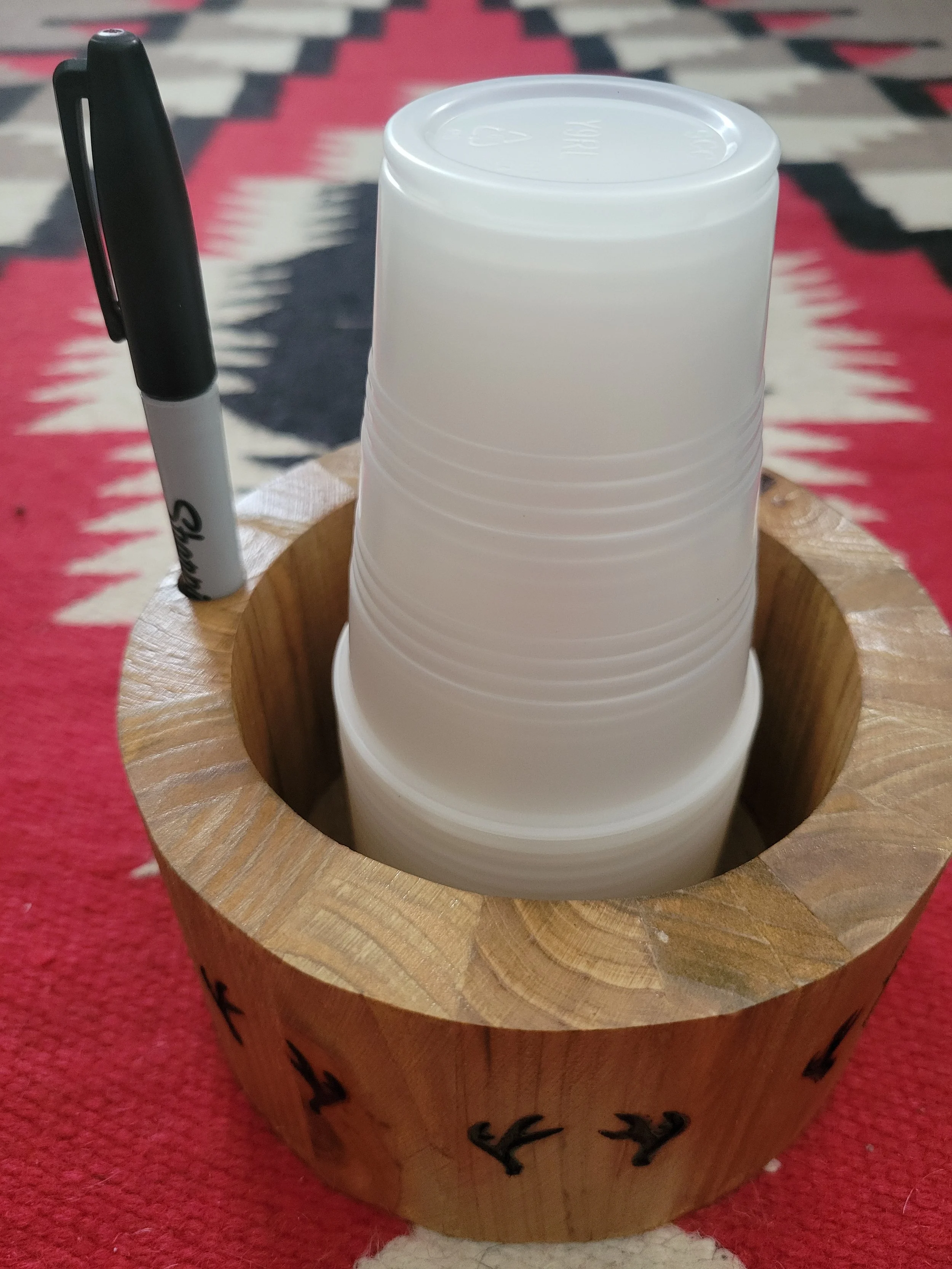 Stacked white plastic cups in a wooden holder with a black marker pen beside it, placed on a patterned rug.