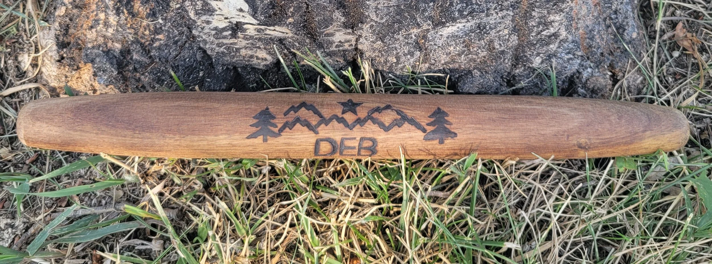 A wooden axe handle with a mountain, tree, and star engraving, and the letters 'DEB' below, lying on grass and soil.