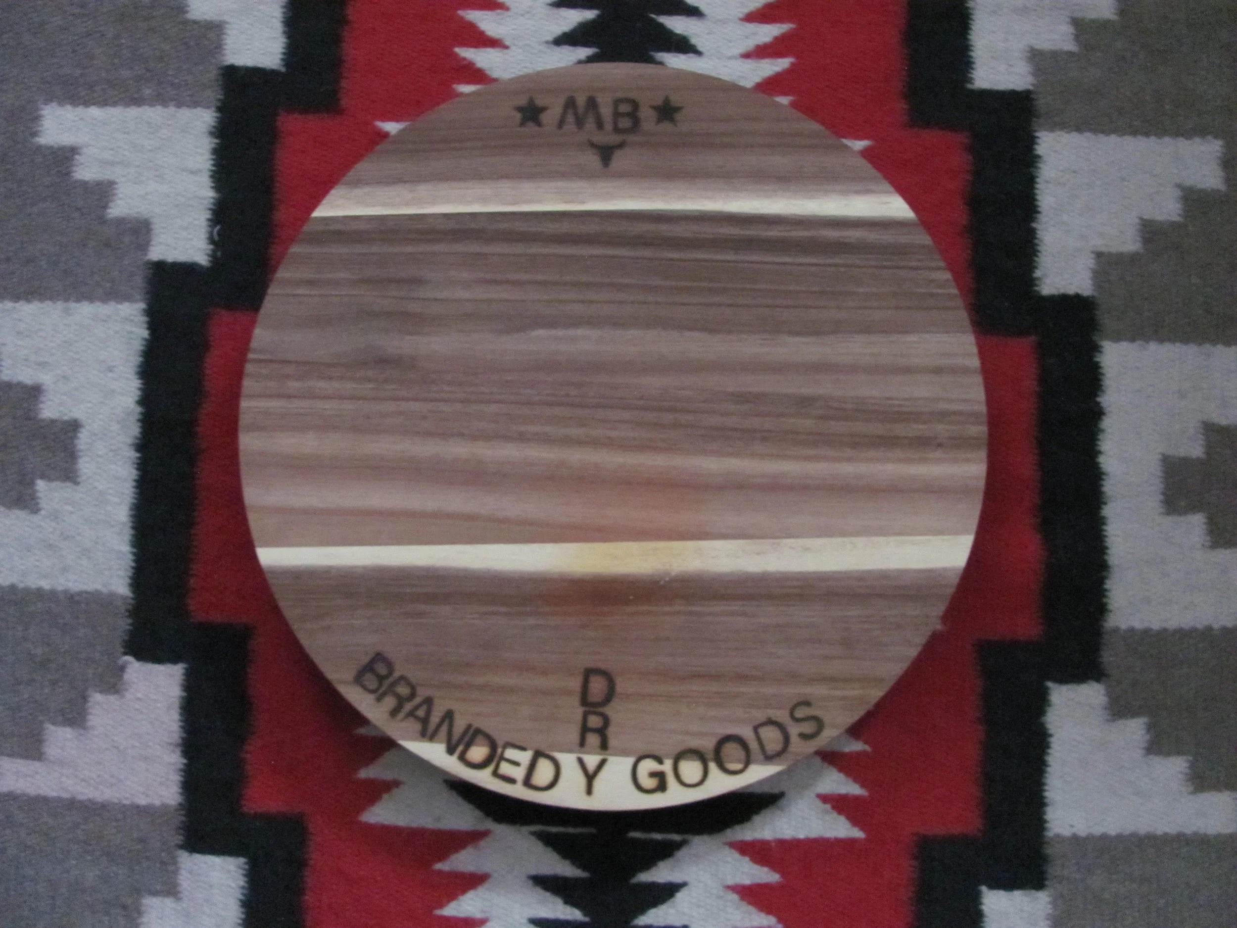 A round wooden cutting board with the words ''BRANDED Y GOODS'' engraved along the bottom edge and ''★ MB ★'' engraved at the top. The cutting board is placed on a patterned rug with geometric designs in red, black, white, and gray.