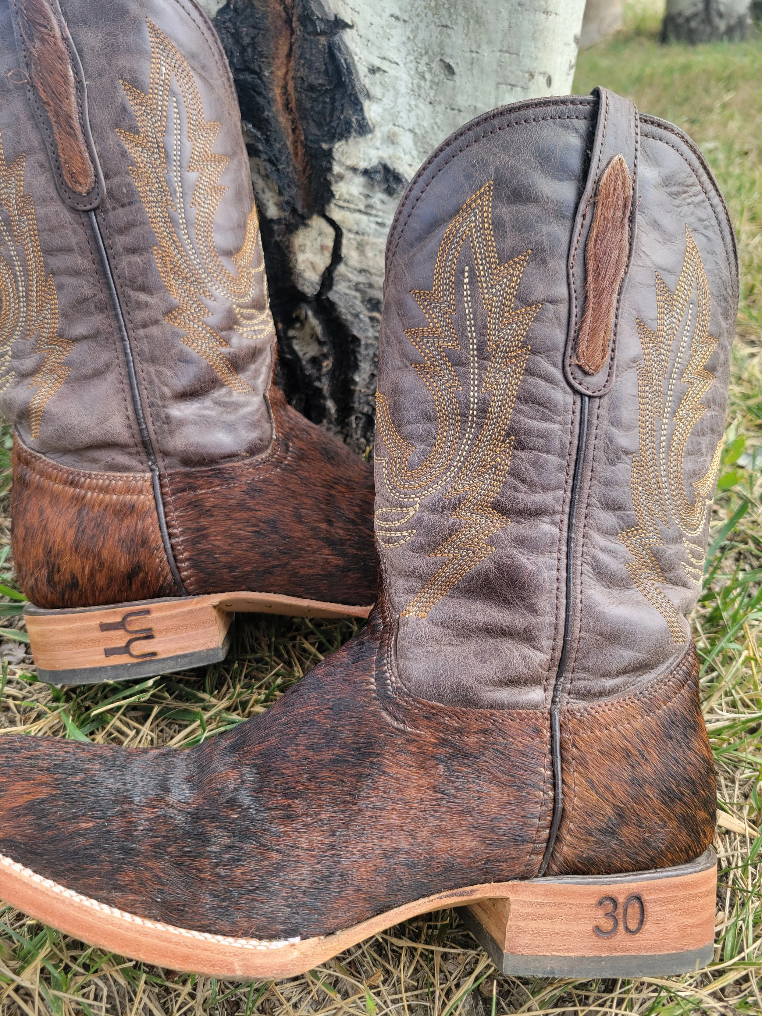 A pair of cowboy boots with a cowhide pattern and embroidered pine tree design, leaning against a tree in a grassy outdoor setting.