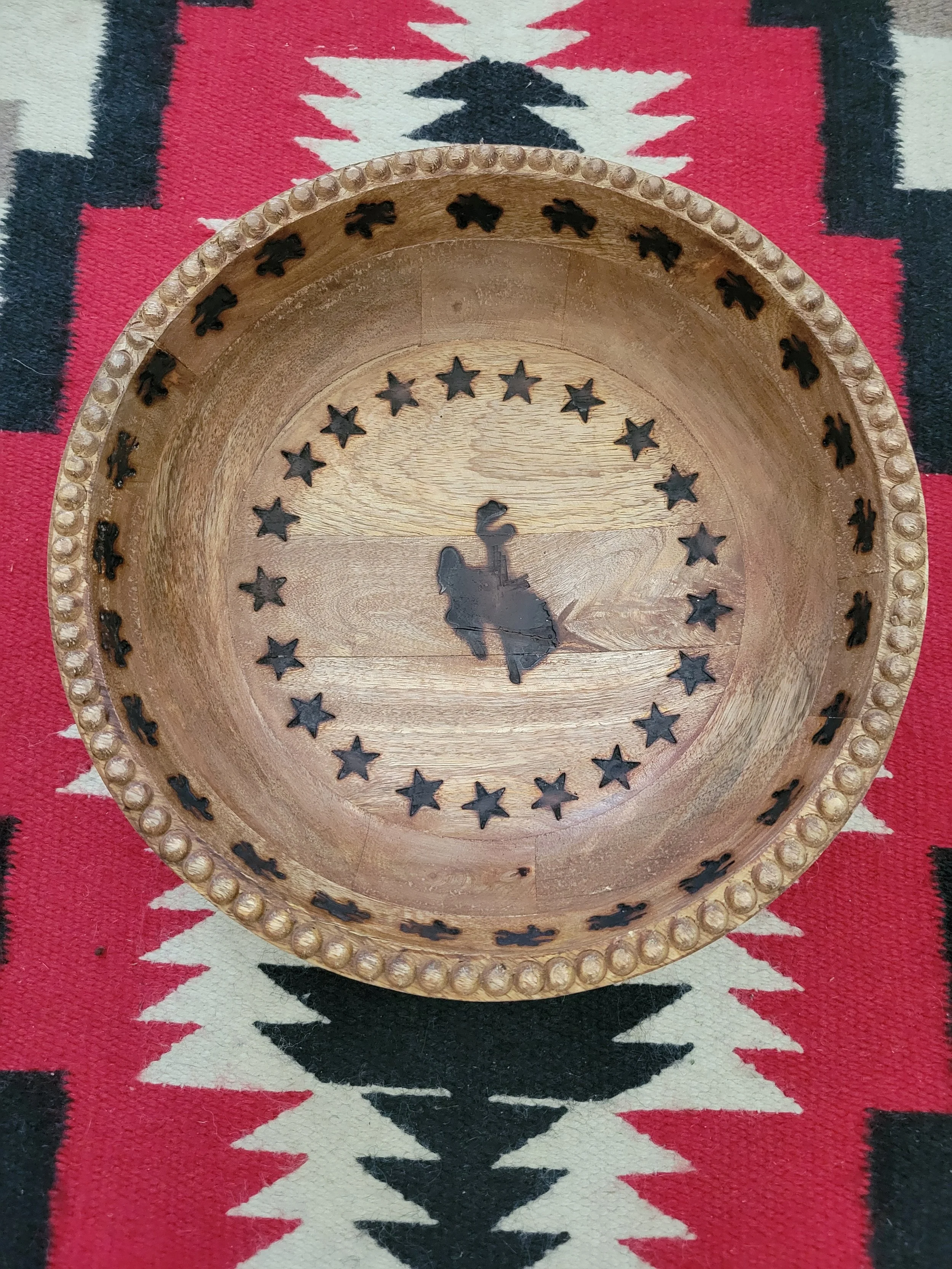 A round wooden tray with a cowboy rider silhouette in the center, surrounded by star-shaped cutouts and beaded edges, placed on a red, black, and white patterned fabric.