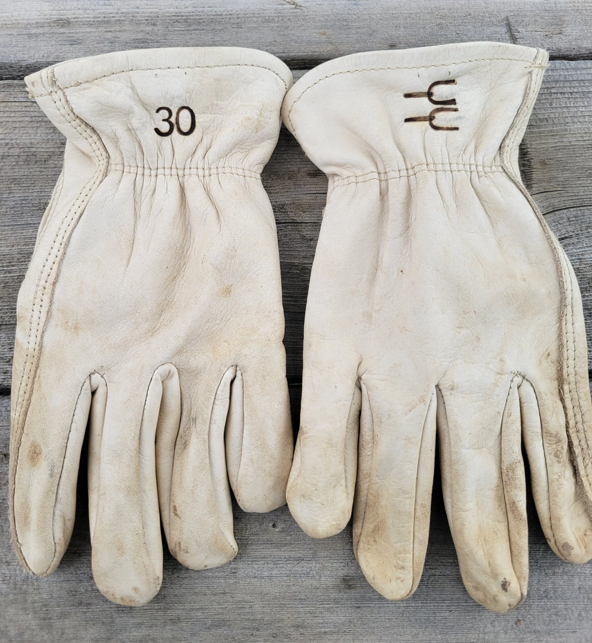 A pair of well-worn vintage cream-colored work gloves with reinforced fingers, one marked with the number 30 on the wrist area.