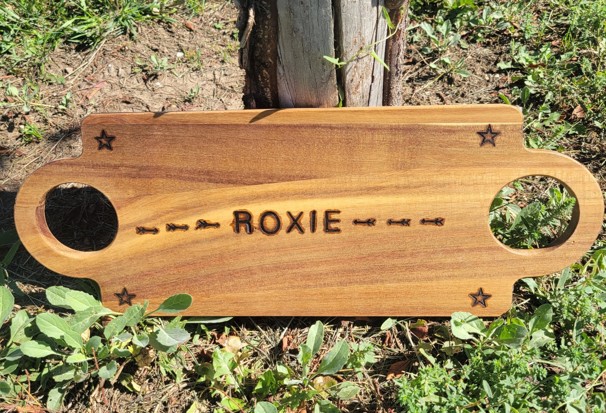 Wooden rectangular sign with rounded corners and holes on each side, placed on soil with green plants. The sign has the name 'Roxie' burned into it and decorative stars at the corners.