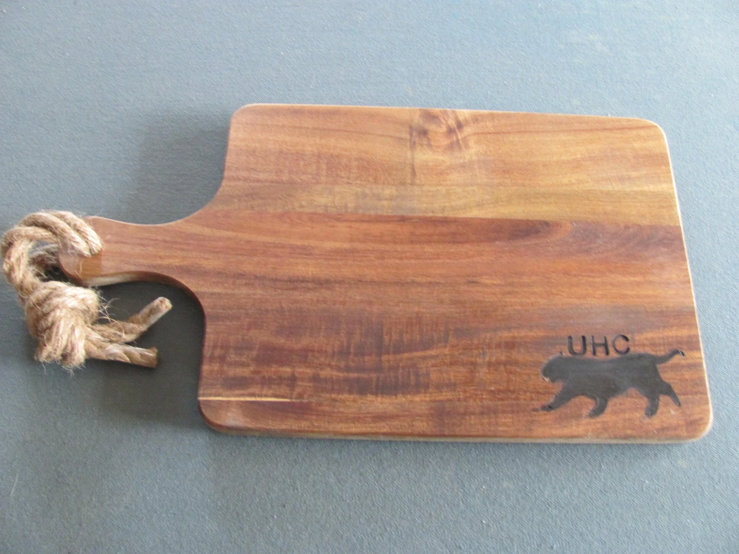 A wooden cutting board with a handle wrapped with a piece of rope, and a silhouette of a bear with the letters 'UHC' engraved on it.