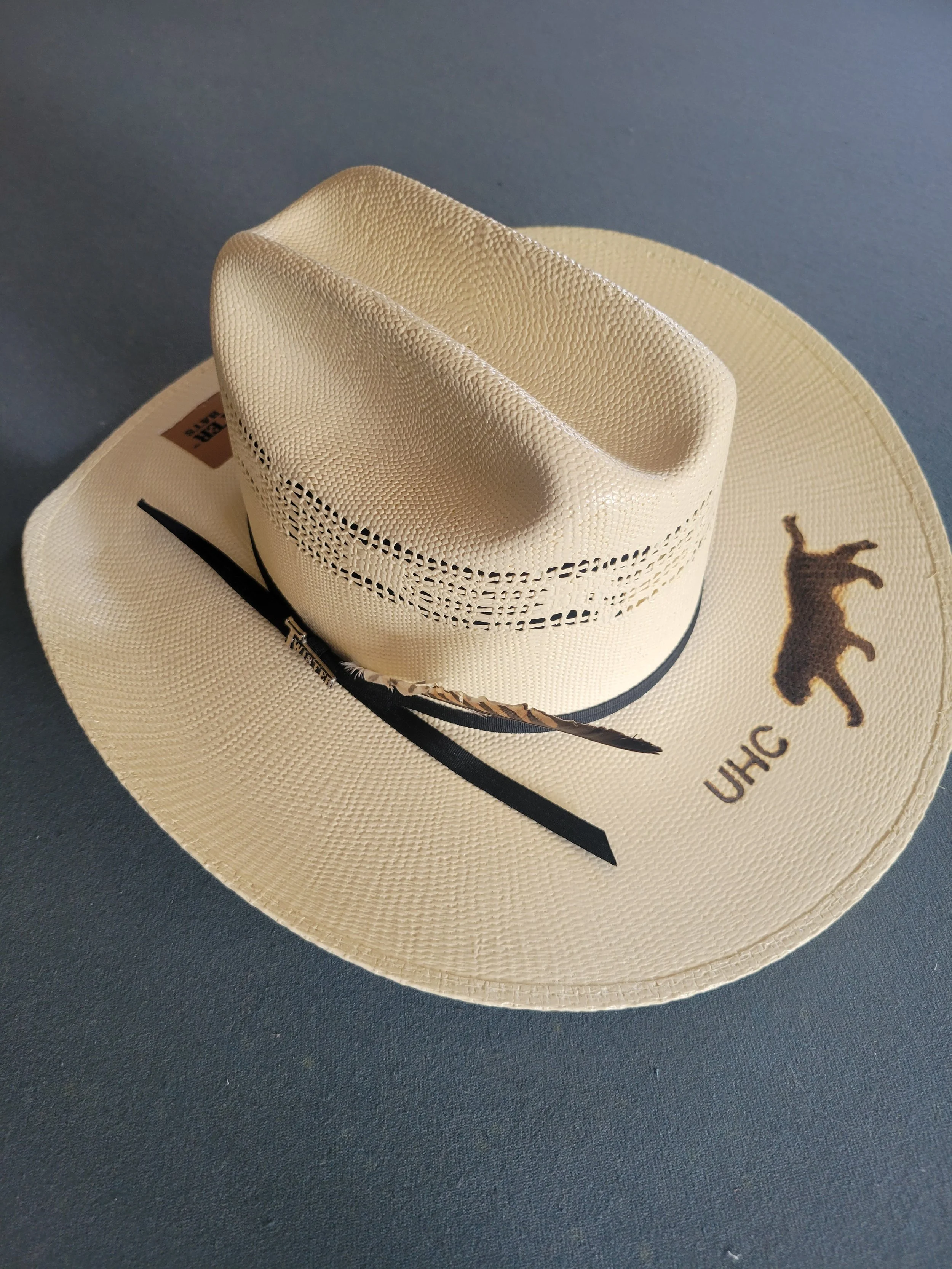Cream-colored cowboy hat with UHC logo, black band, and decorative feather, placed on a dark surface.