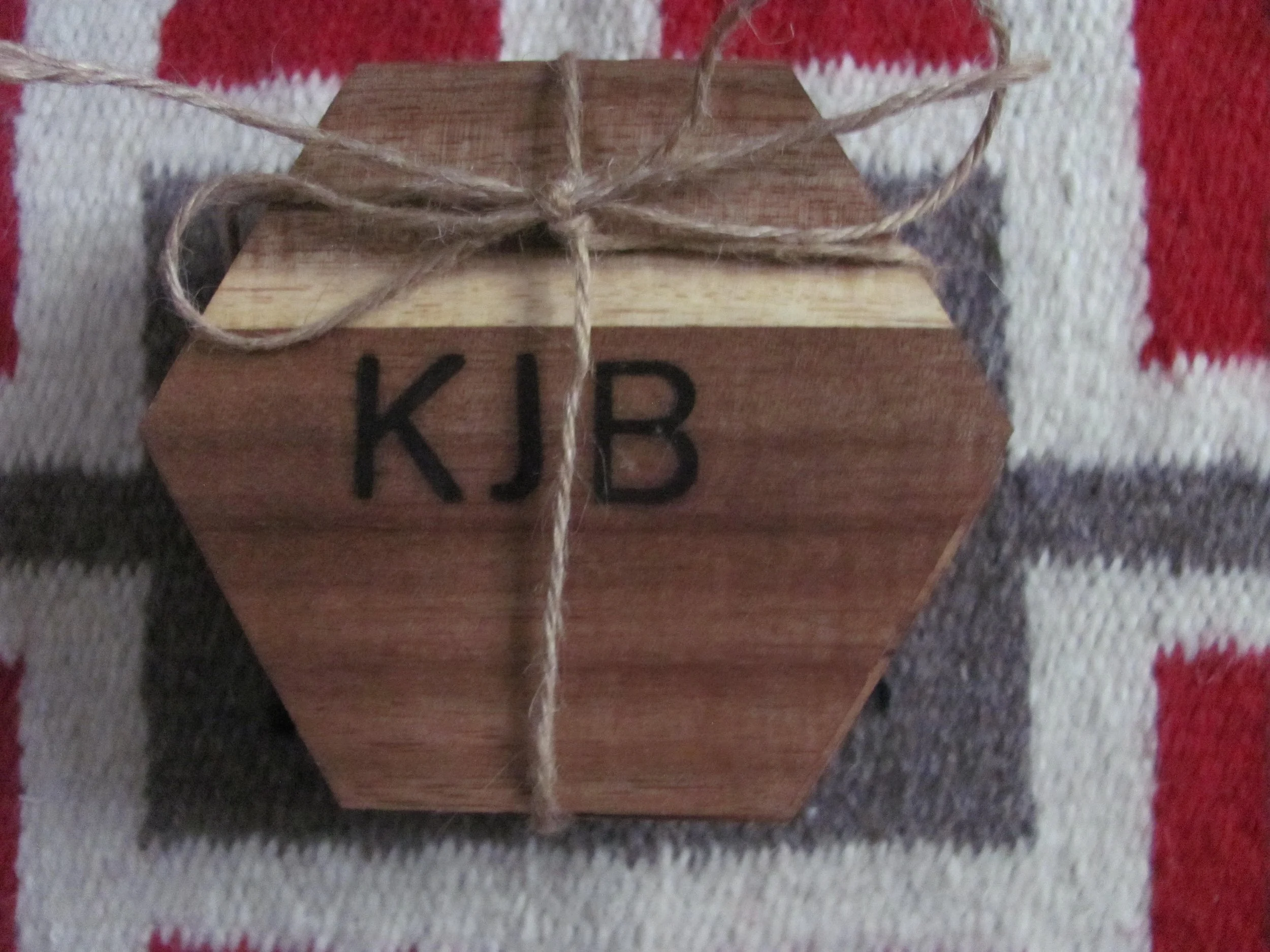A wooden cube with the initials 'KJB' painted in black on one side, tied with a piece of string, resting on a patterned fabric surface.