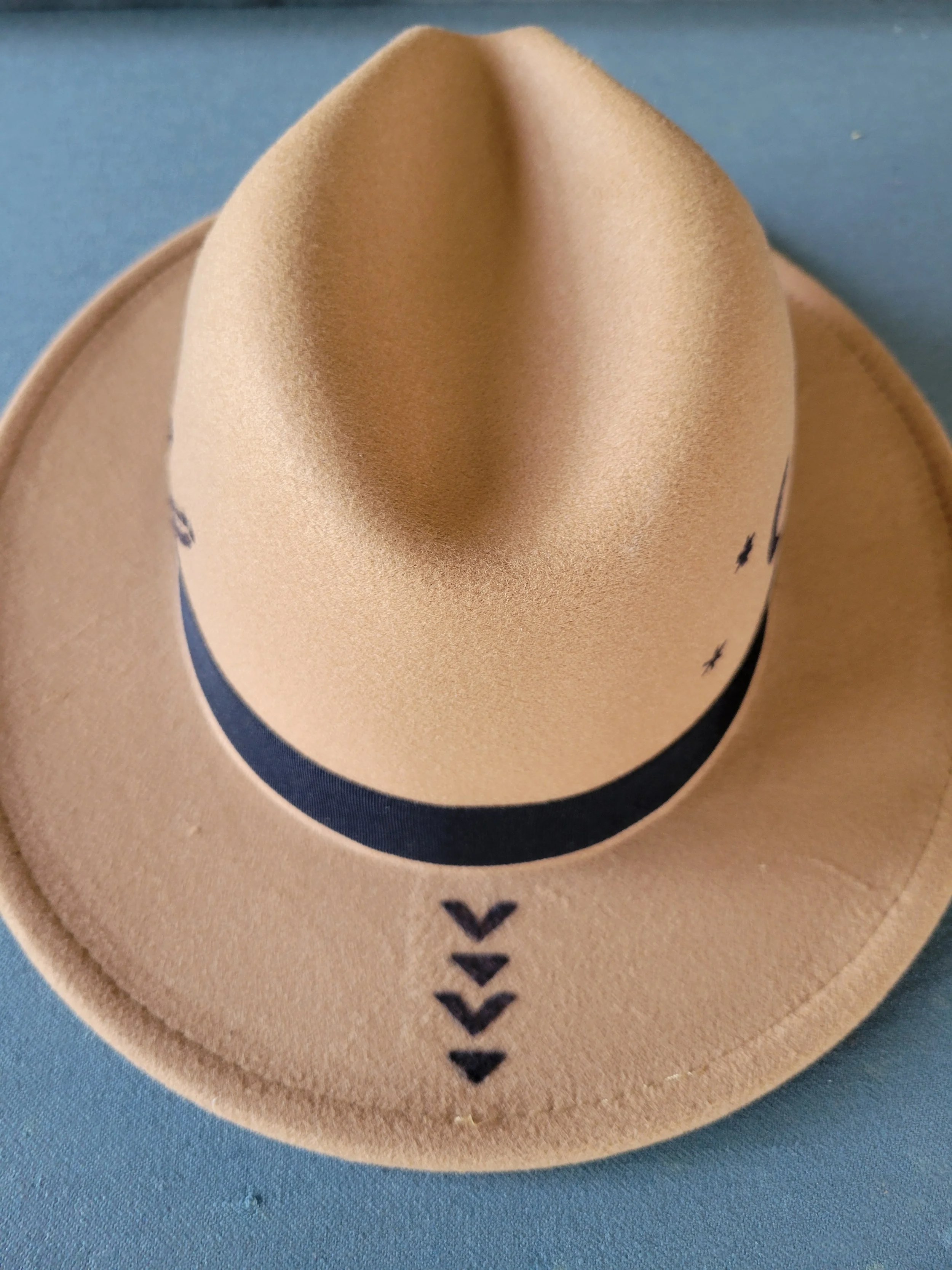 A beige cowboy hat with black decorative designs on the brim, placed on a blue surface.