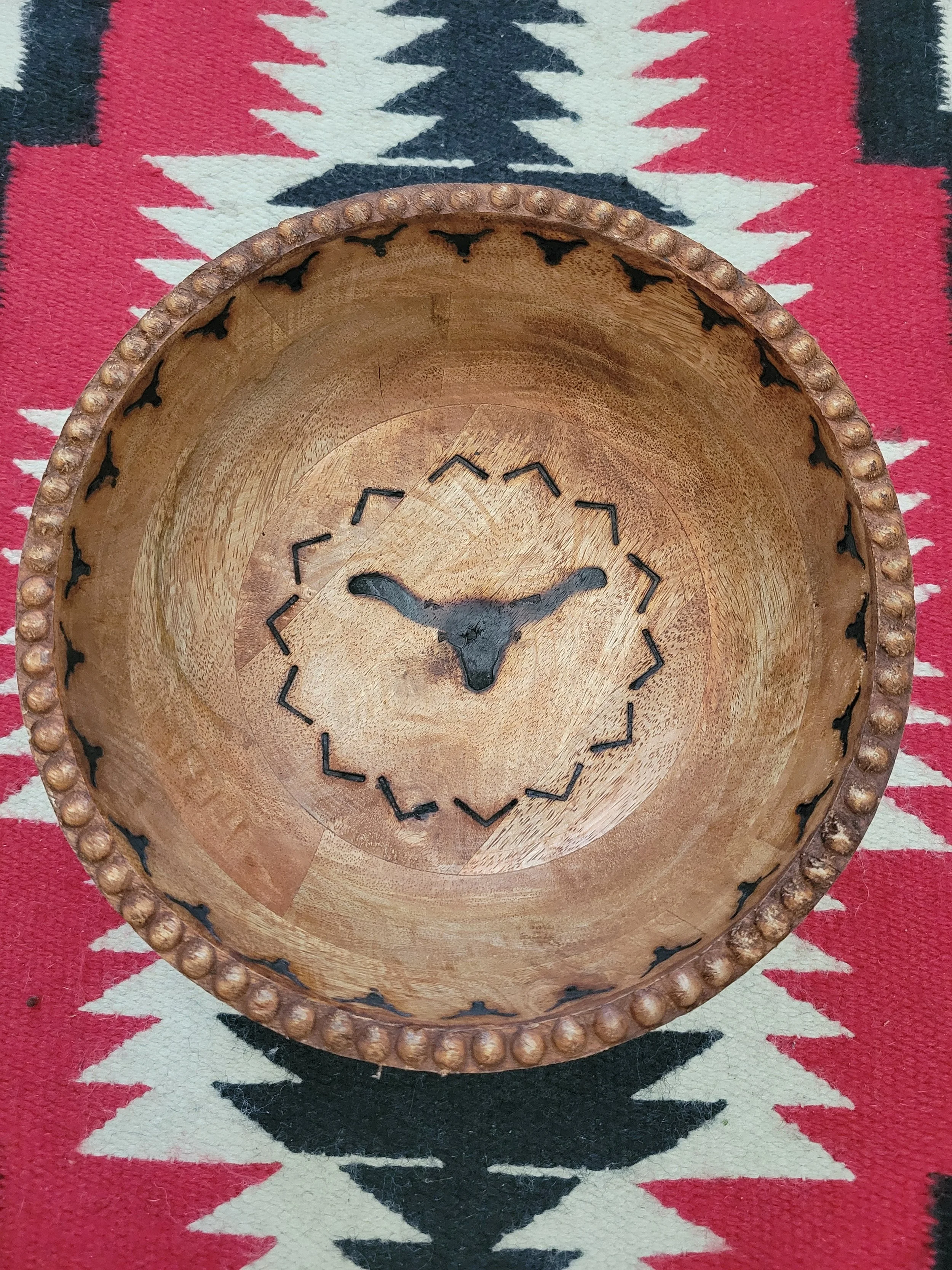 A round wooden tray with a decorative beaded edge, featuring a silhouette of a longhorn steer head in the center, placed on a patterned textile with red, black, and white geometric designs.