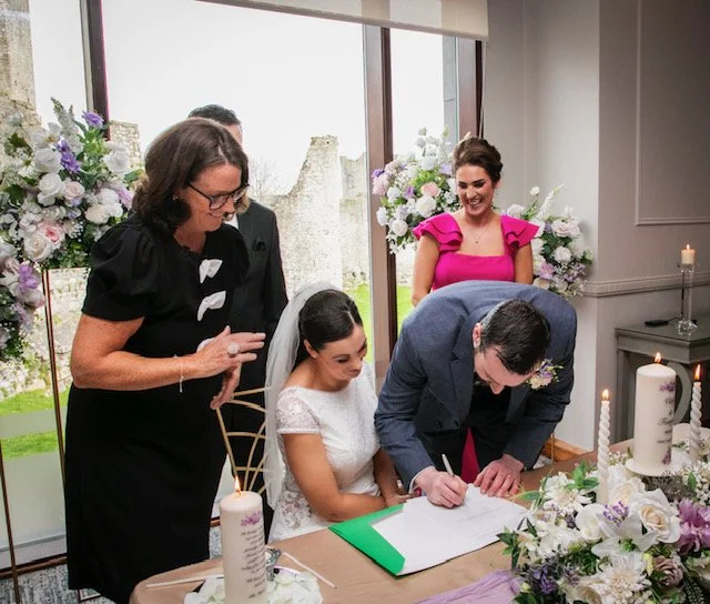 A wedding ceremony with a bride and groom signing a marriage register at a table decorated with flowers and candles, with guests overseeing the process.