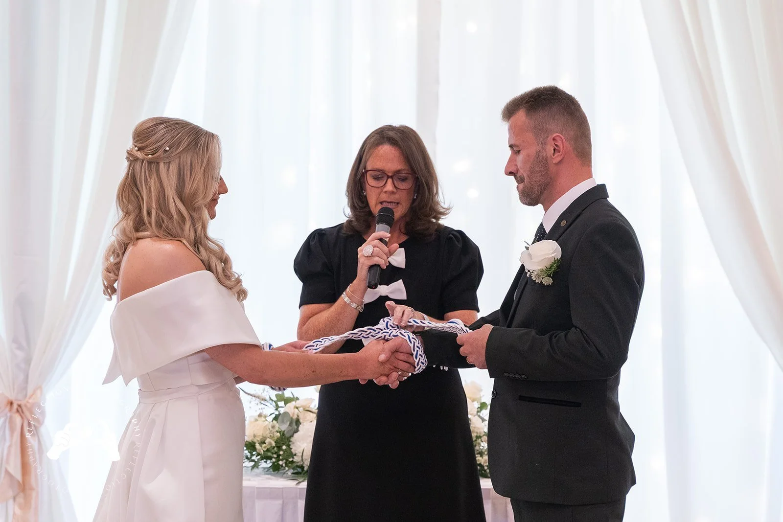 A wedding ceremony with a bride and groom holding hands and exchanging vows, officiated by a woman holding a microphone, against a backdrop of white curtains and floral decorations.