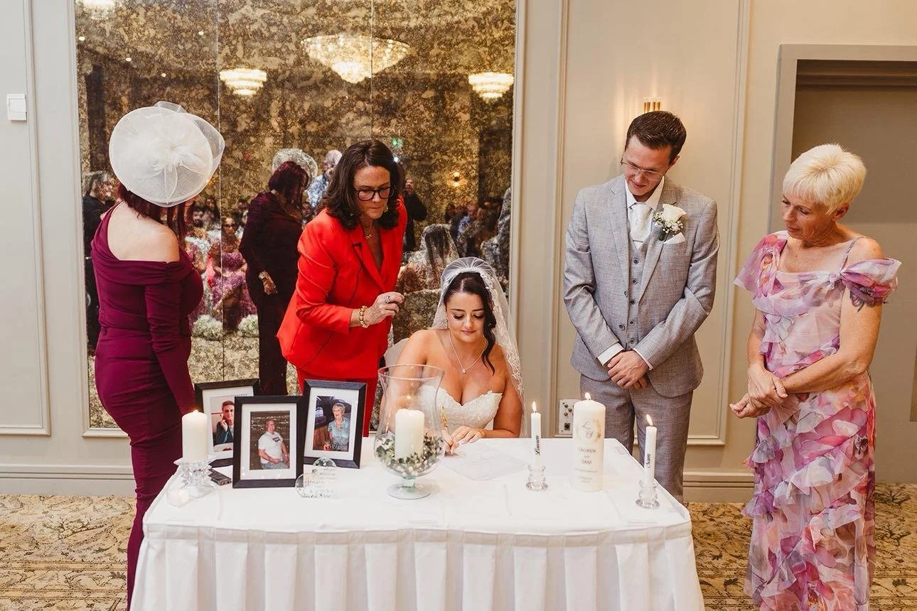 Bride seated at a table signing a document during a wedding ceremony, surrounded by family members in elegant attire, with framed photos, candles, and a glass vase on the table.