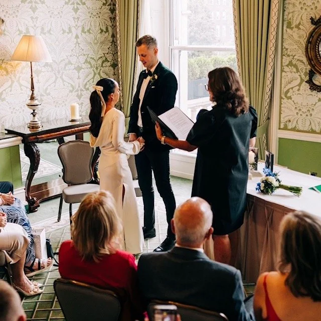 A wedding ceremony with a girl and a man holding hands, facing each other, in front of an officiant, during the daytime indoors with natural light. Guests seated and watching attentively.