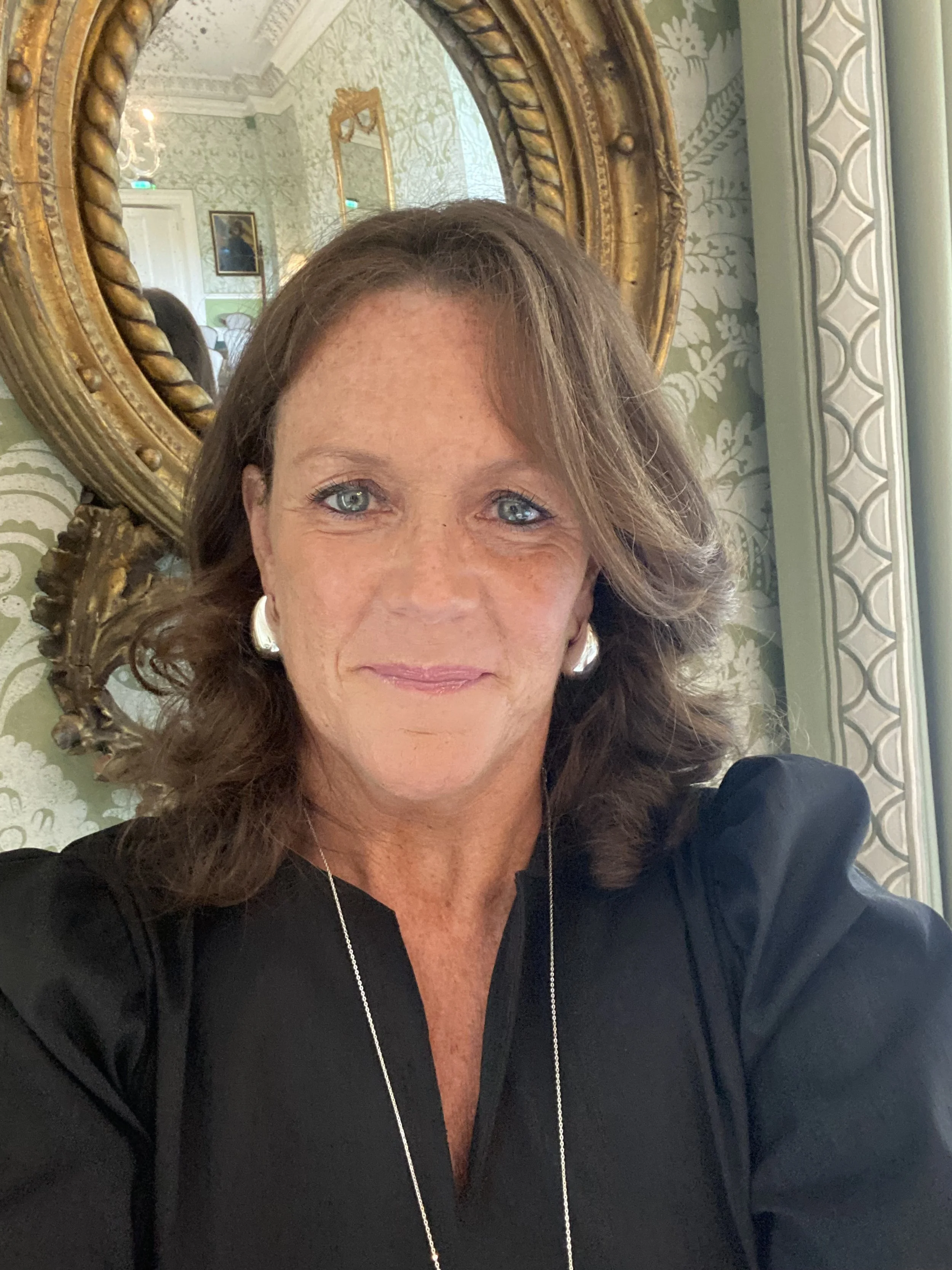 A woman with wavy brown hair and blue eyes taking a selfie in front of an ornate gold framed mirror, with elegant wallpaper and a chandelier visible in the background.