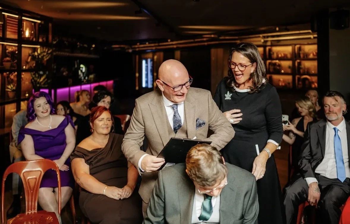 A group of people in formal attire at an indoor event, with two women and one man smiling and conversing, while others seated around them look on.