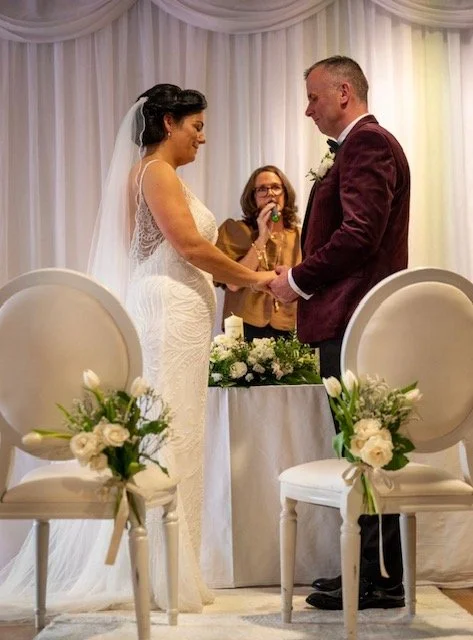 Bride and groom holding hands during wedding ceremony, standing in front of officiant, with floral arrangements on white chairs in the foreground.