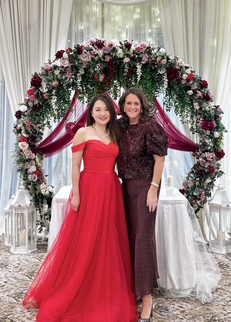 Two women standing together in front of a floral arch, smiling. One woman is wearing a red gown, and the other is wearing a dark purple outfit. The setting appears to be a decorated indoor venue, possibly for a celebration or wedding.