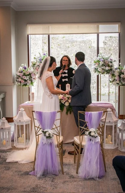 Bride and groom holding hands during their wedding ceremony in front of an officiant, with floral arrangements and lanterns in the background.