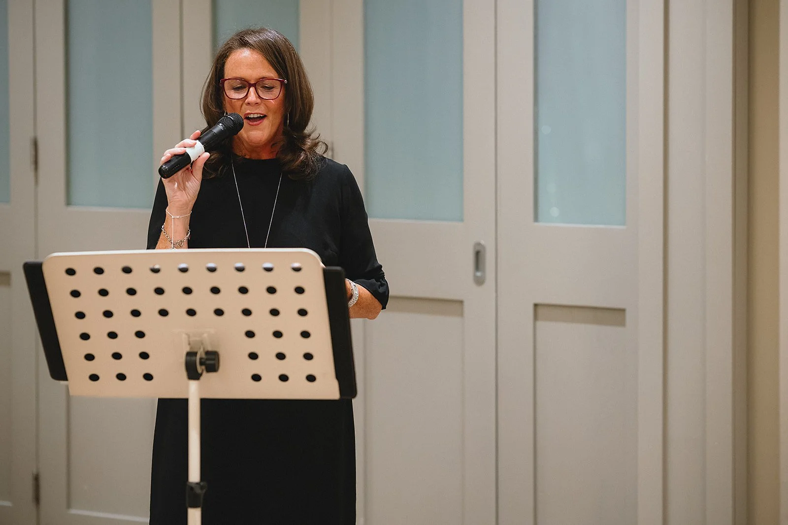 A woman with brown hair, red glasses, and wearing a black dress is singing into a microphone. She is standing behind a music stand in a room with closed wooden doors and frosted glass panels.