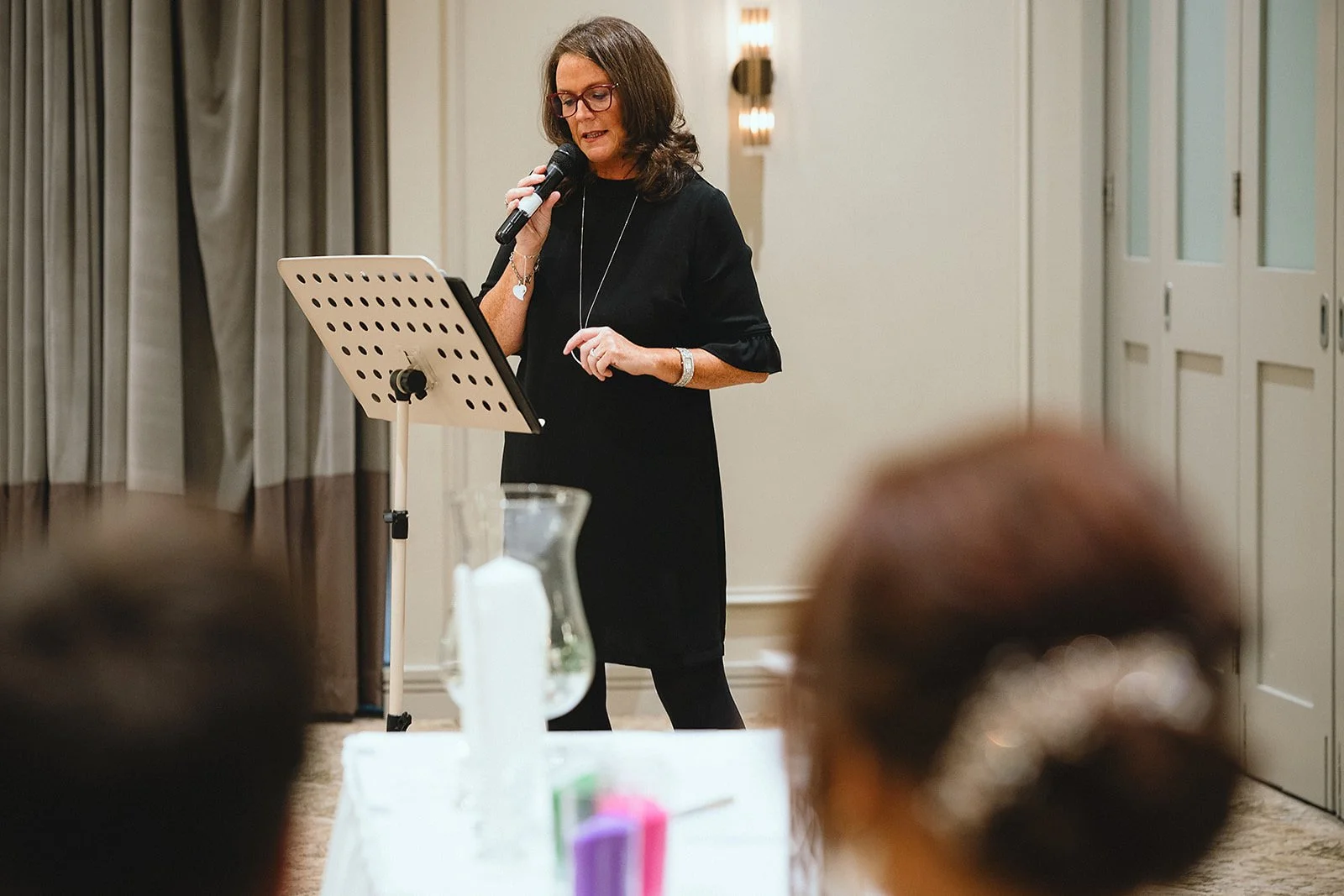 Woman in black dress wearing glasses, speaking into a microphone at a lectern in a conference room.