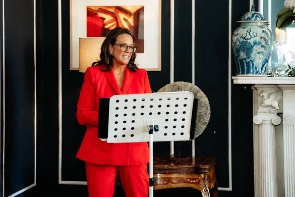 A woman in a bright red suit speaking or singing behind a music stand in a decorated room.