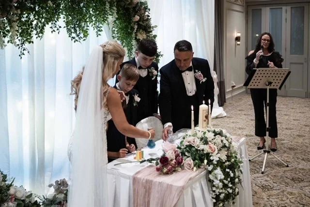 A wedding ceremony with a bride and groom signing a document, surrounded by two children, with an officiant and a musician in the background.