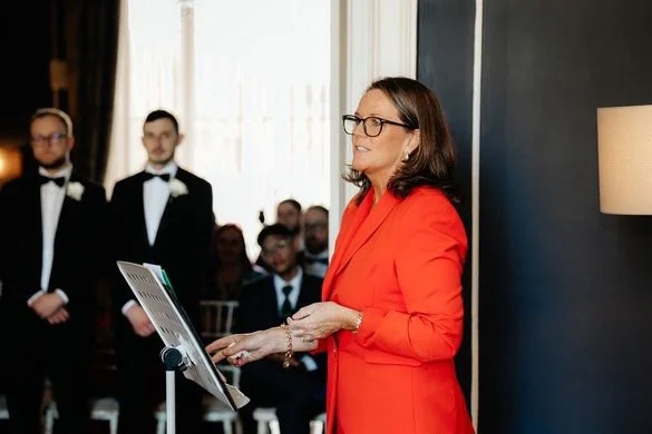 A woman in a red blazer giving a speech or presentation in front of an audience at an indoor event.